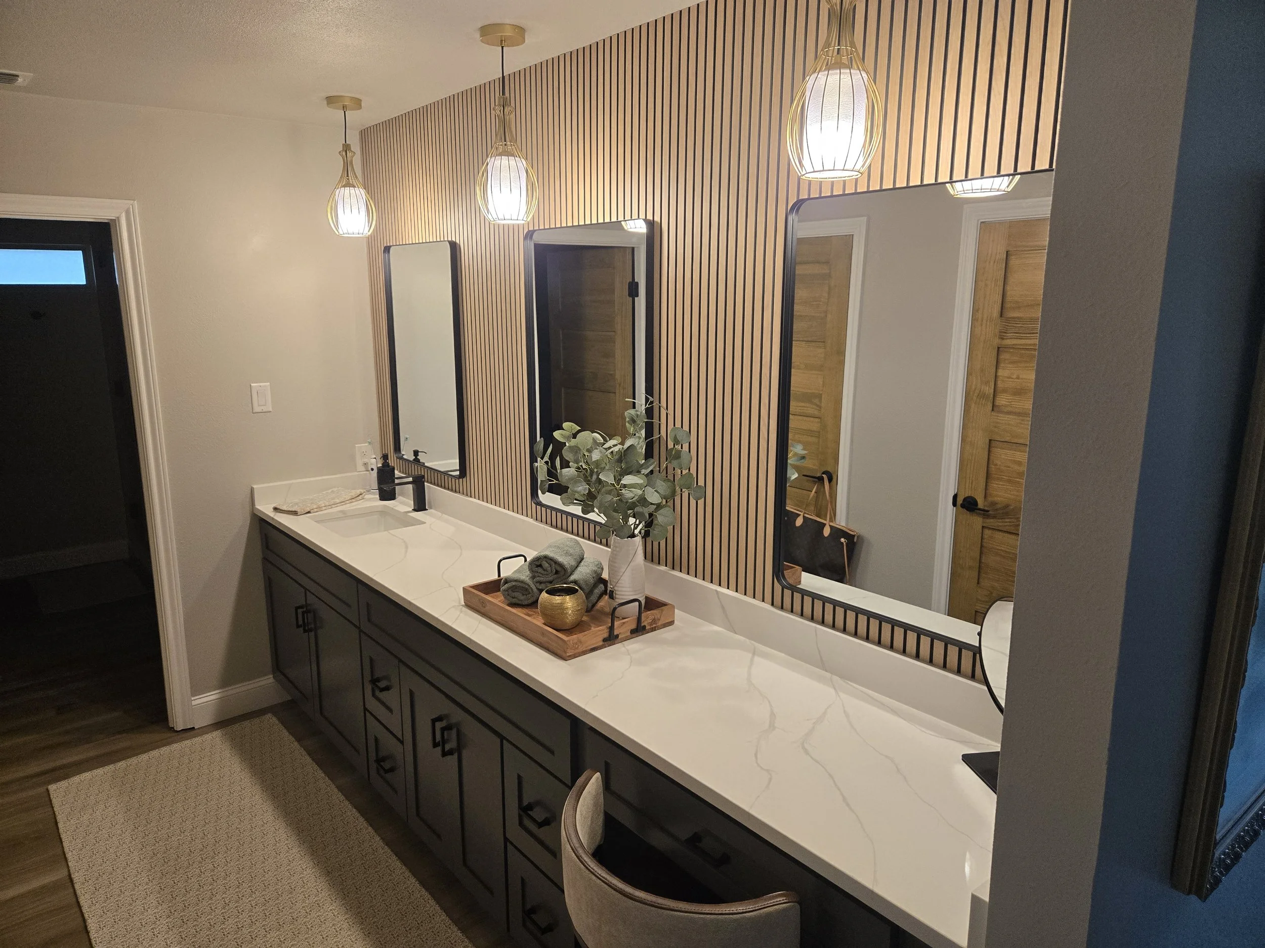 A bathroom vanity with a white countertop, black cabinets, and a mirror framed with black. Above the mirror, three pendant lights hang from the ceiling. On the counter, there is a green plant in a white vase, a tray with rolled towels, and a soap dispenser.