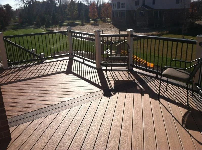 Sunlit wooden deck with a metal chair and a bench, surrounded by a black metal railing, overlooking a grassy backyard with trees and a house in the background.