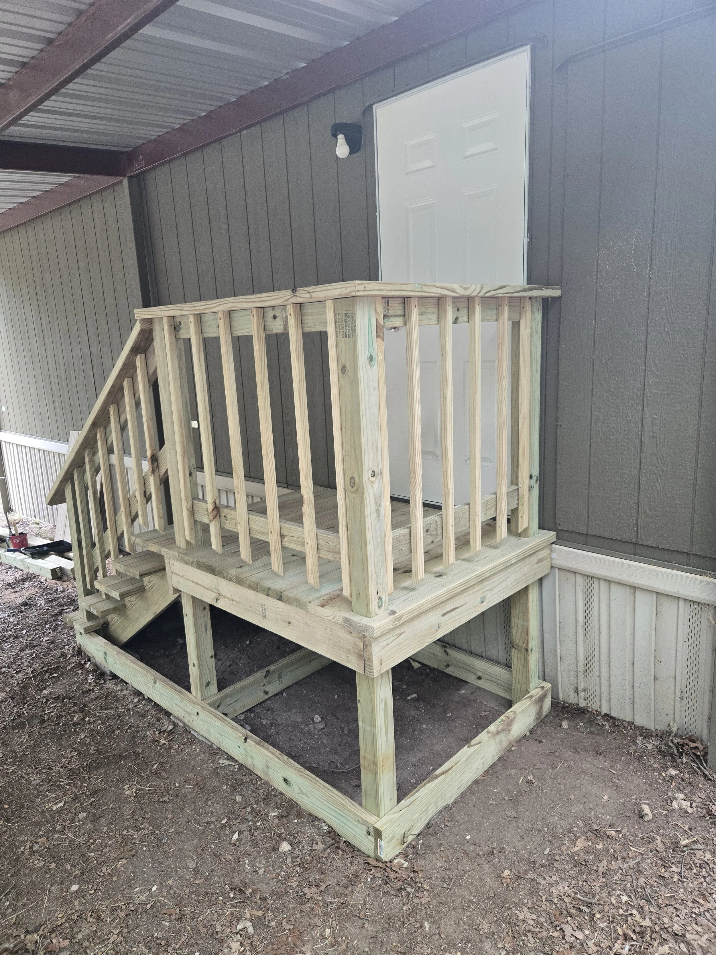 Unfinished wooden deck with railing, attached to the side of a house with gray paneling, under a metal roof.