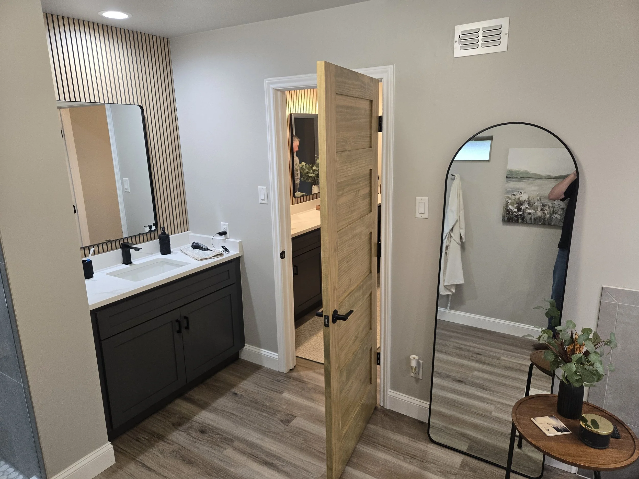A modern bathroom with a white vanity, black cabinet, large mirror, striped accent wall, open wooden door leading to a room with a dresser, wall art, and person taking a photo in a full-length mirror. There are small tables with plants and decor on the right side.