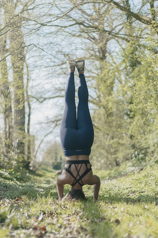 femme faisant la pose de poirier dans une forêt, entourée d'arbres et de verdure, sous un ciel ensoleillé.