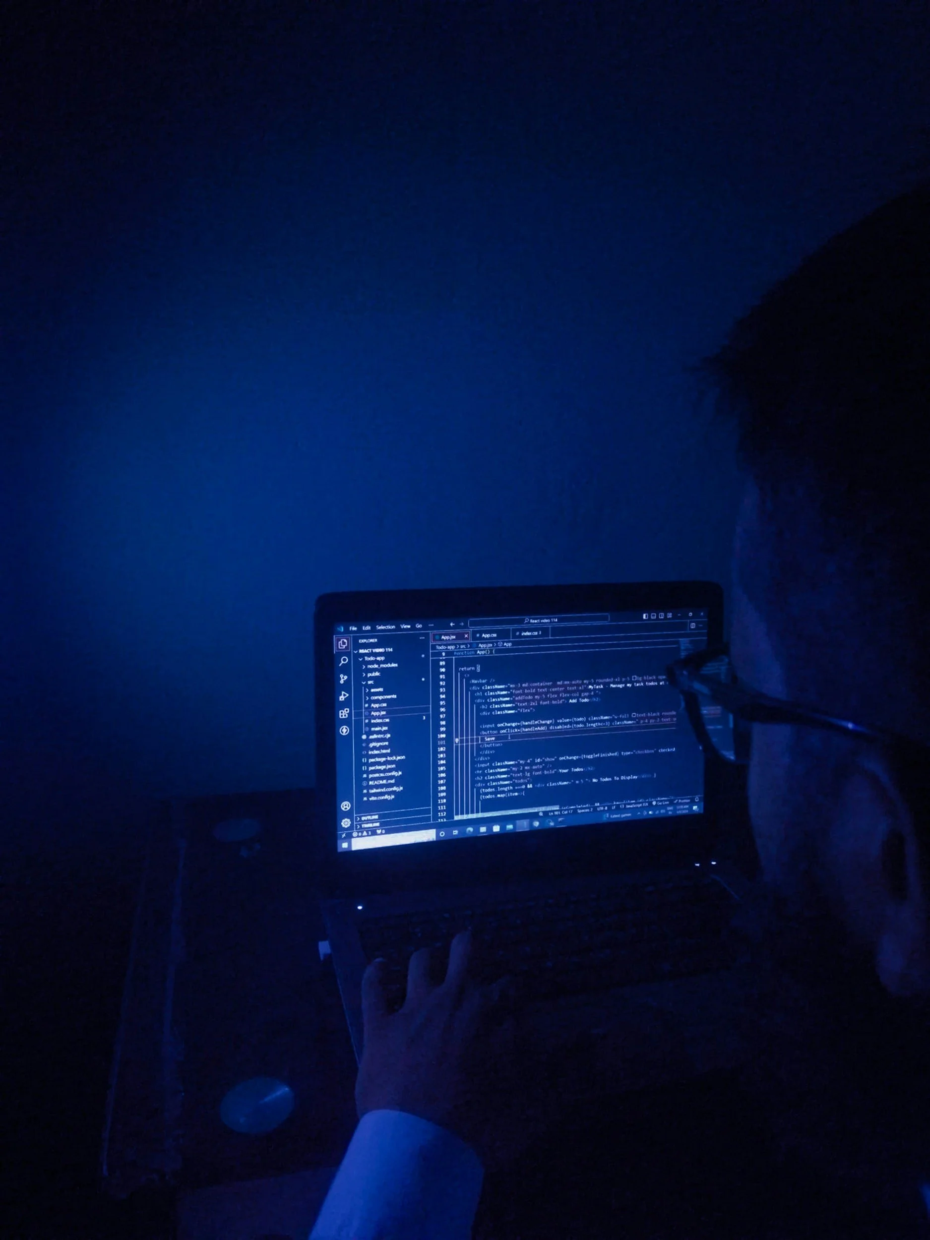 Dark room with a computer monitor displaying code, a person's leg in shorts, and a hand pointing at the screen.