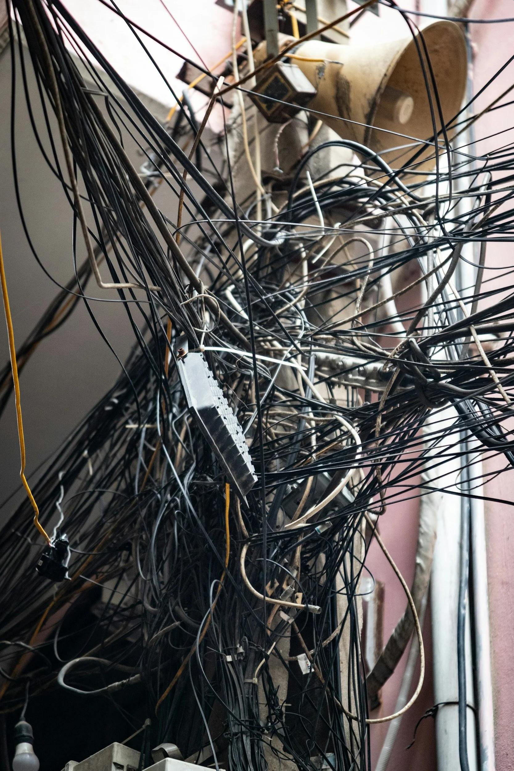 A tangled mess of numerous black and white electrical wires and cables hanging on a wall, with some electronic components and connectors visible among the cables.