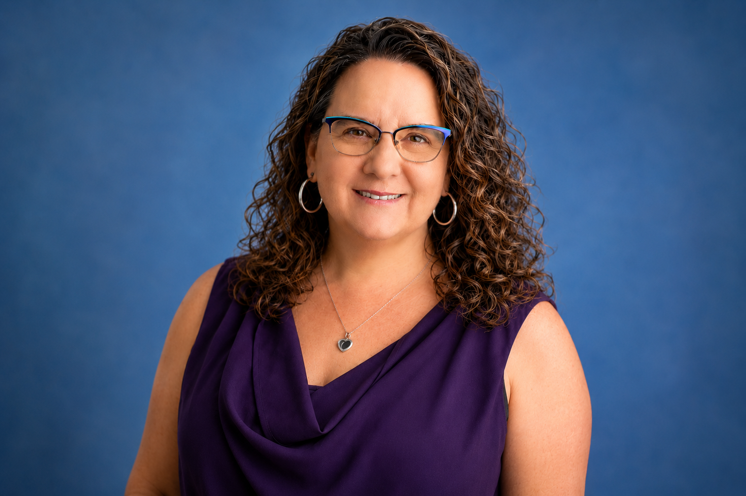 A woman with curly brown hair wearing glasses, hoop earrings, a heart-shaped necklace, and a sleeveless purple dress, smiling against a blue background.