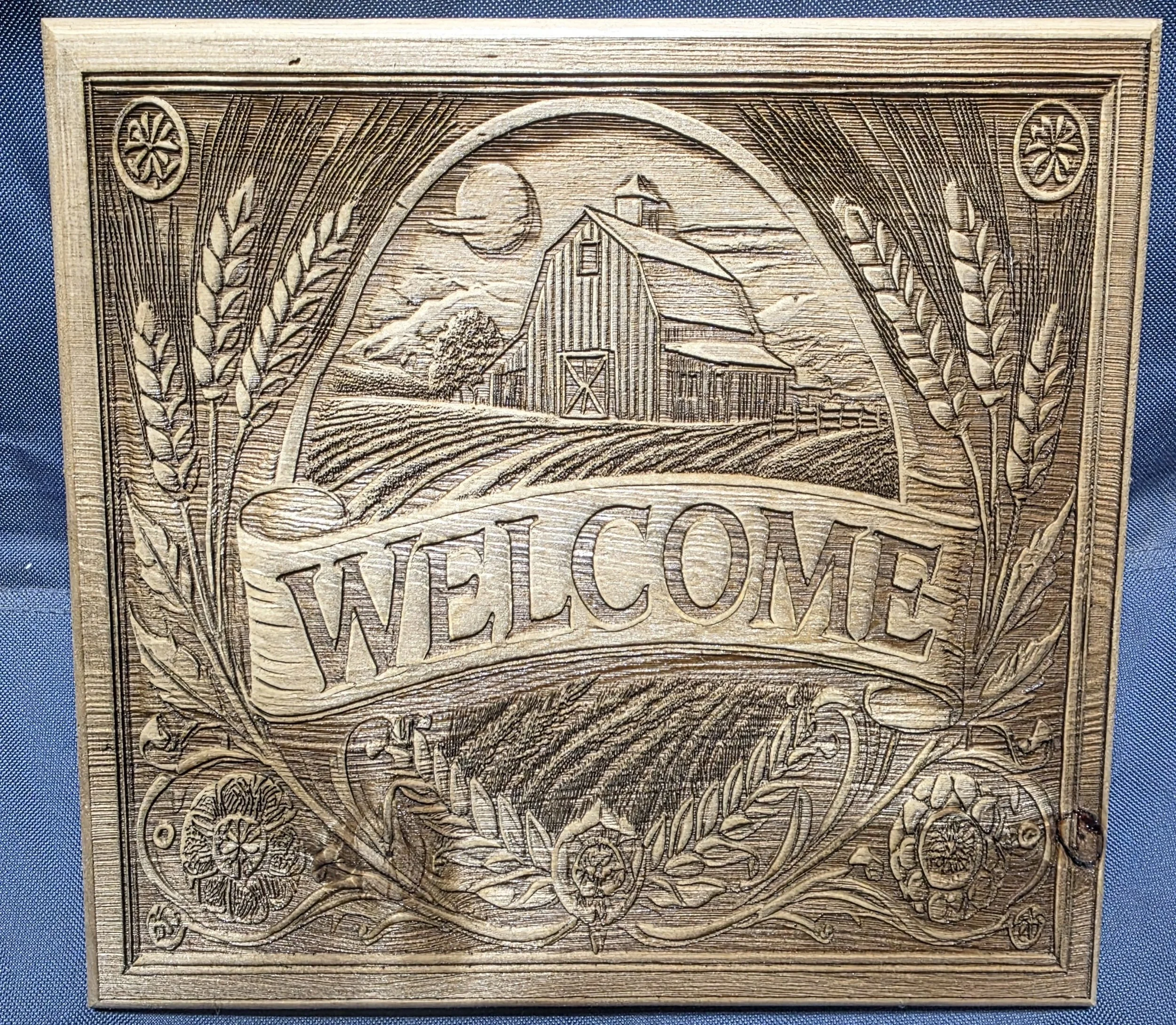 Wooden carved plaque with detailed illustrations of a barn, mountains, the sun, and wheat stalks surrounding the scene.