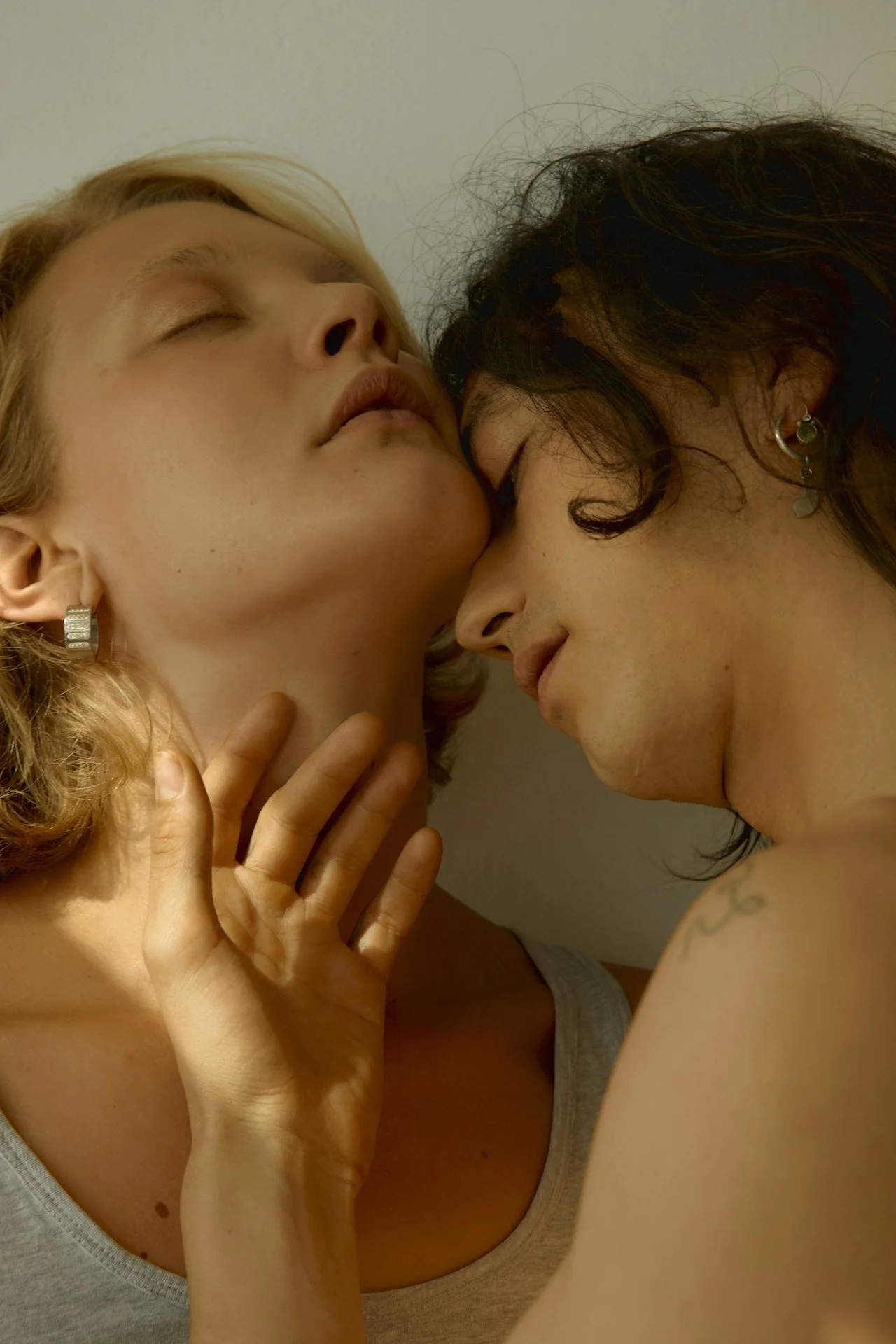 Two women with their foreheads touching, eyes closed, and gentle expressions. One has blonde hair and wears earrings, while the other has darker, curly hair and a tattoo on her arm.