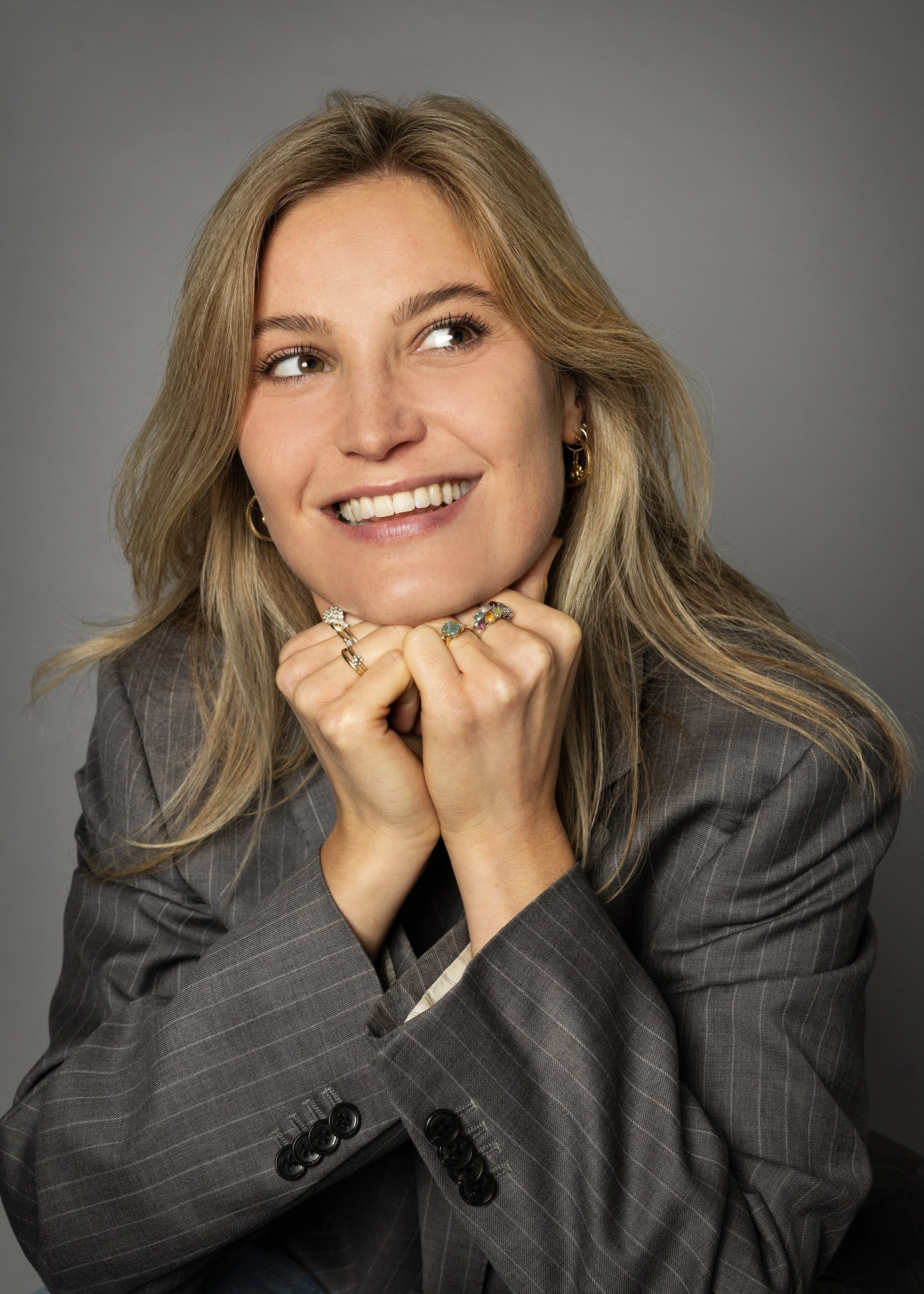 Charlotte Sun glimlachend met lang, golvend blond haar, gekleed in een grijs, gestreept blazer, die haar kin met beide handen steunt, gelegen tegen een effen grijze achtergrond.