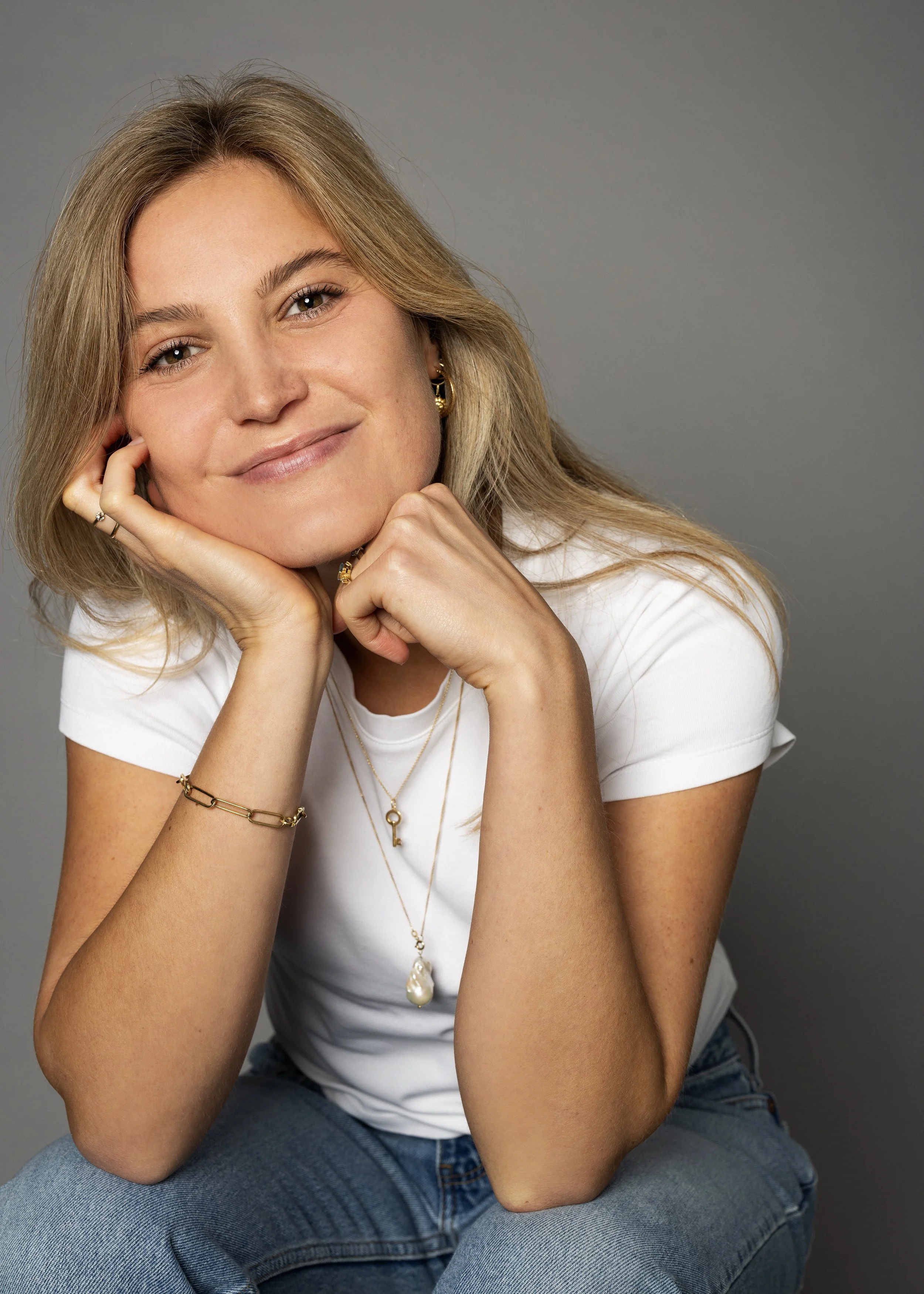 Portret van Charlotte Sun met blond haar die glimlacht, rust haar hoofd op haar hand, draagt gouden sieraden en een witte T-shirt, zittend op een grijze achtergrond.