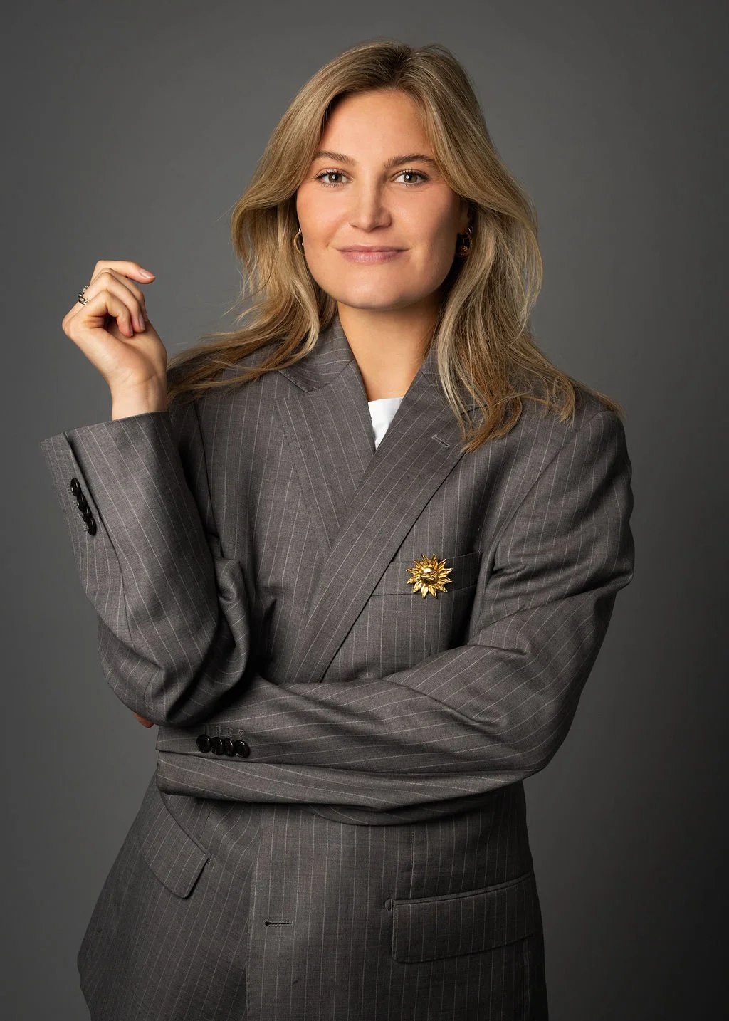 Charlotte Sun in een grijs pinstripe pak met een gouden broche, poseert tegen een effen grijze achtergrond.