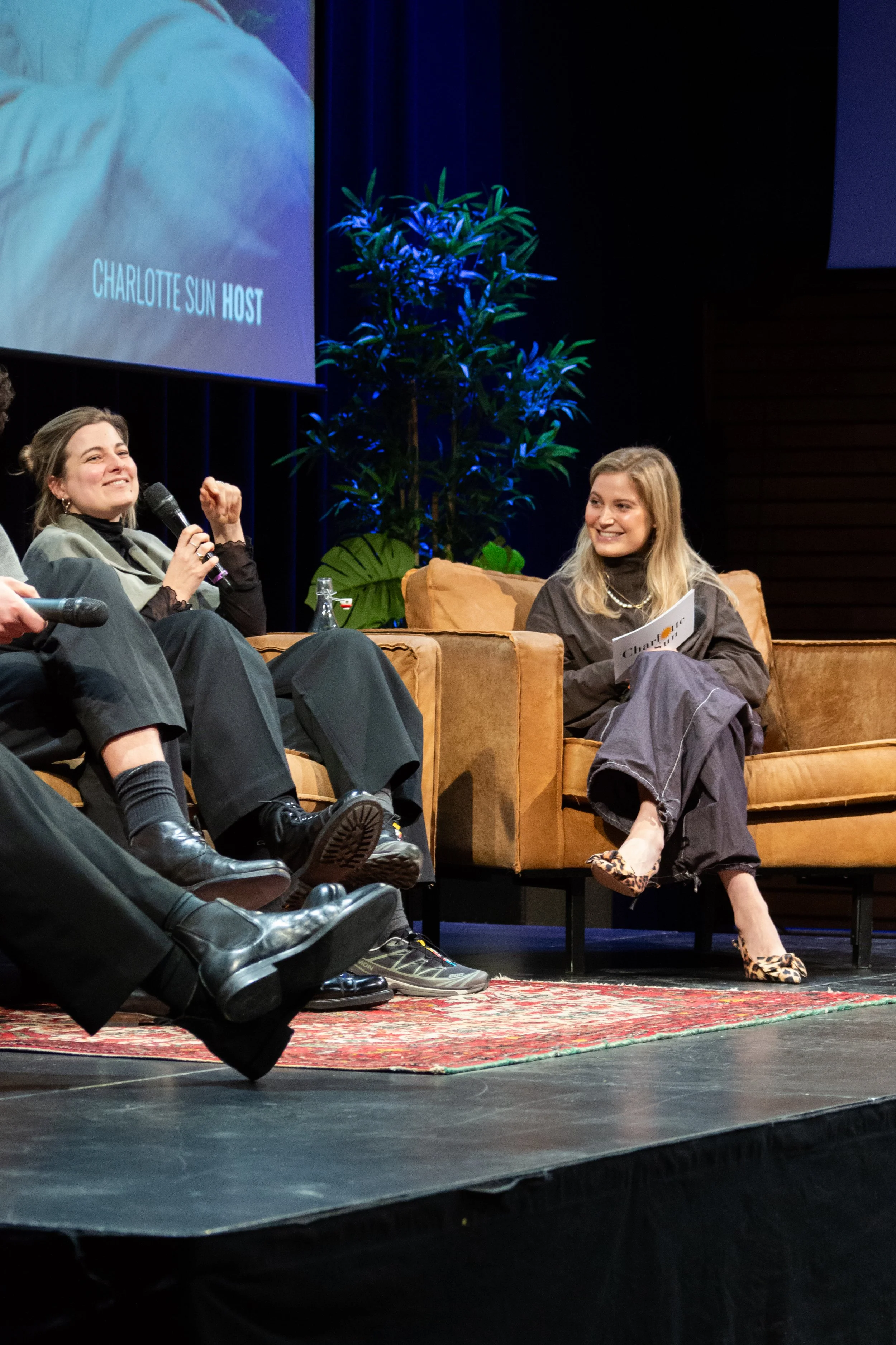 Charlotte Sun m op een podiumpodium met een andere vrouw tijdens een panelgesprek, één houdt een microfoon vast en de ander glimlacht, met een grote planten en een scherm met tekst 'Charlotte Sun Host' in de achtergrond.
