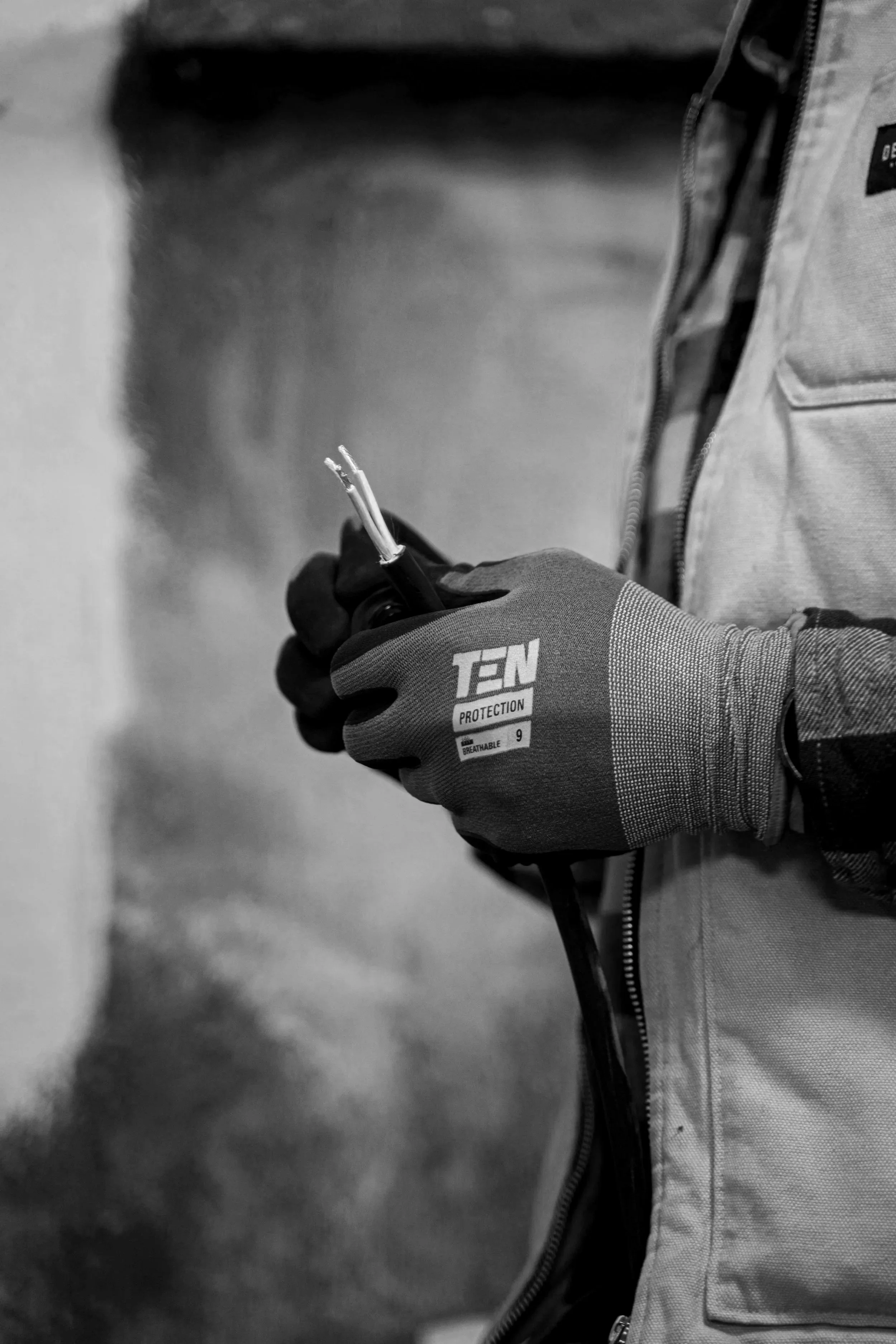 Close-up of a person's hand holding a cable with frayed wires. The person is wearing a glove labeled 'TEN PROTECTION.' They are dressed in a jacket with a zipper and a pocket.