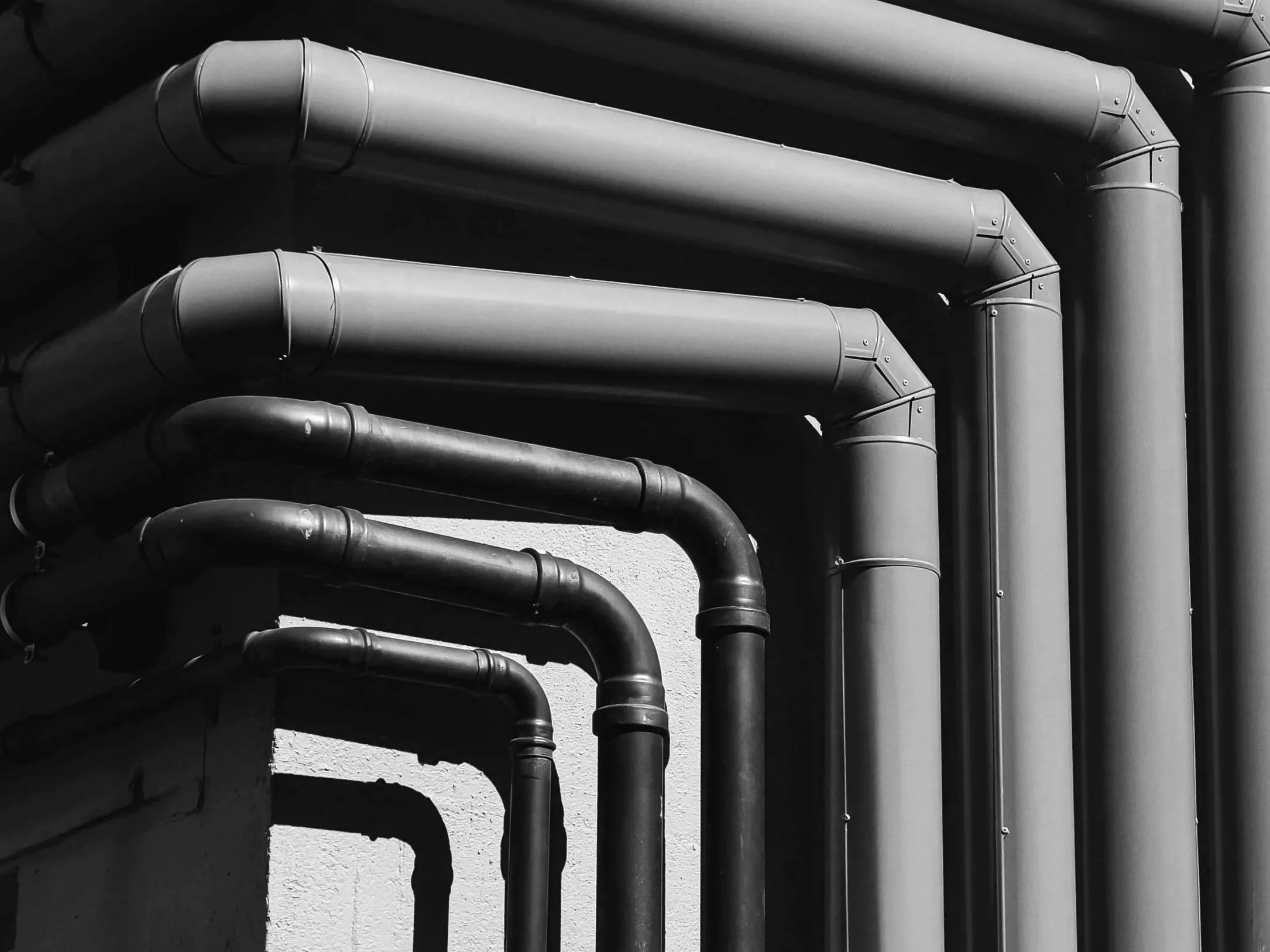 Black and white photo of industrial pipes running along a wall, casting shadows.