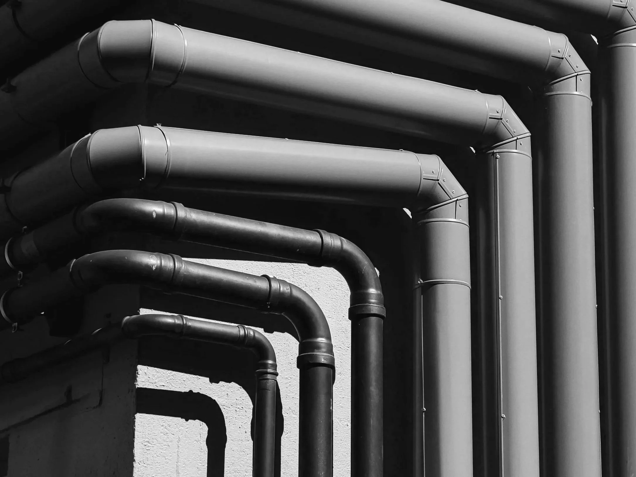 Black and white image of several industrial pipes mounted on a wall, running horizontally across the image with some shadows cast on the wall.