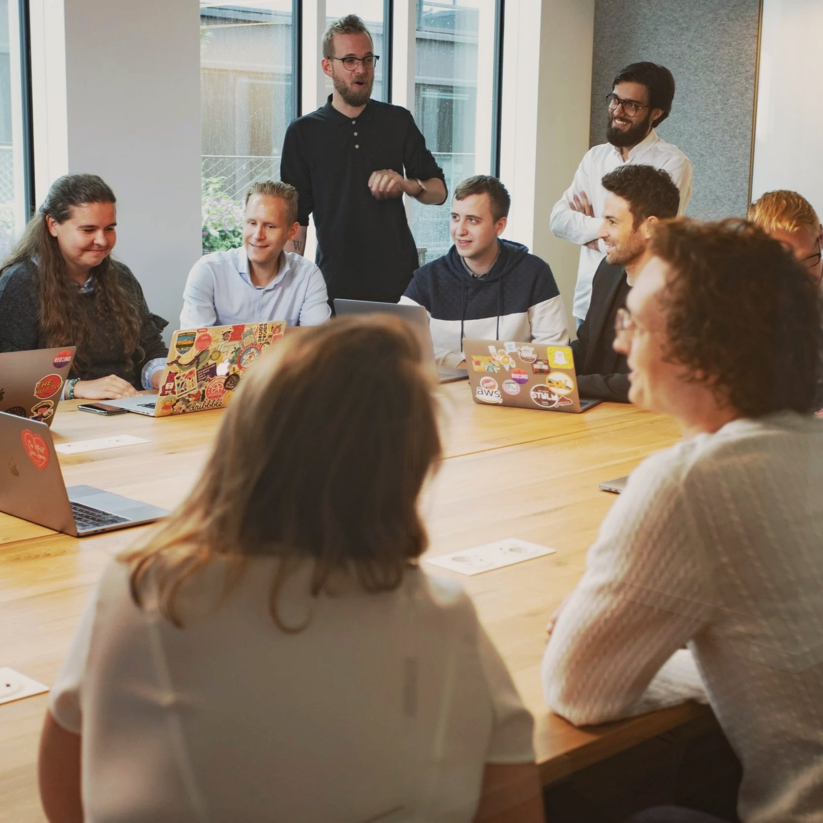 Groupe de personnes assises autour d'une table en réunion, un homme se tenant debout semble parler ou présenter, tous souriants. La scène se déroule dans une salle avec de grandes fenêtres.