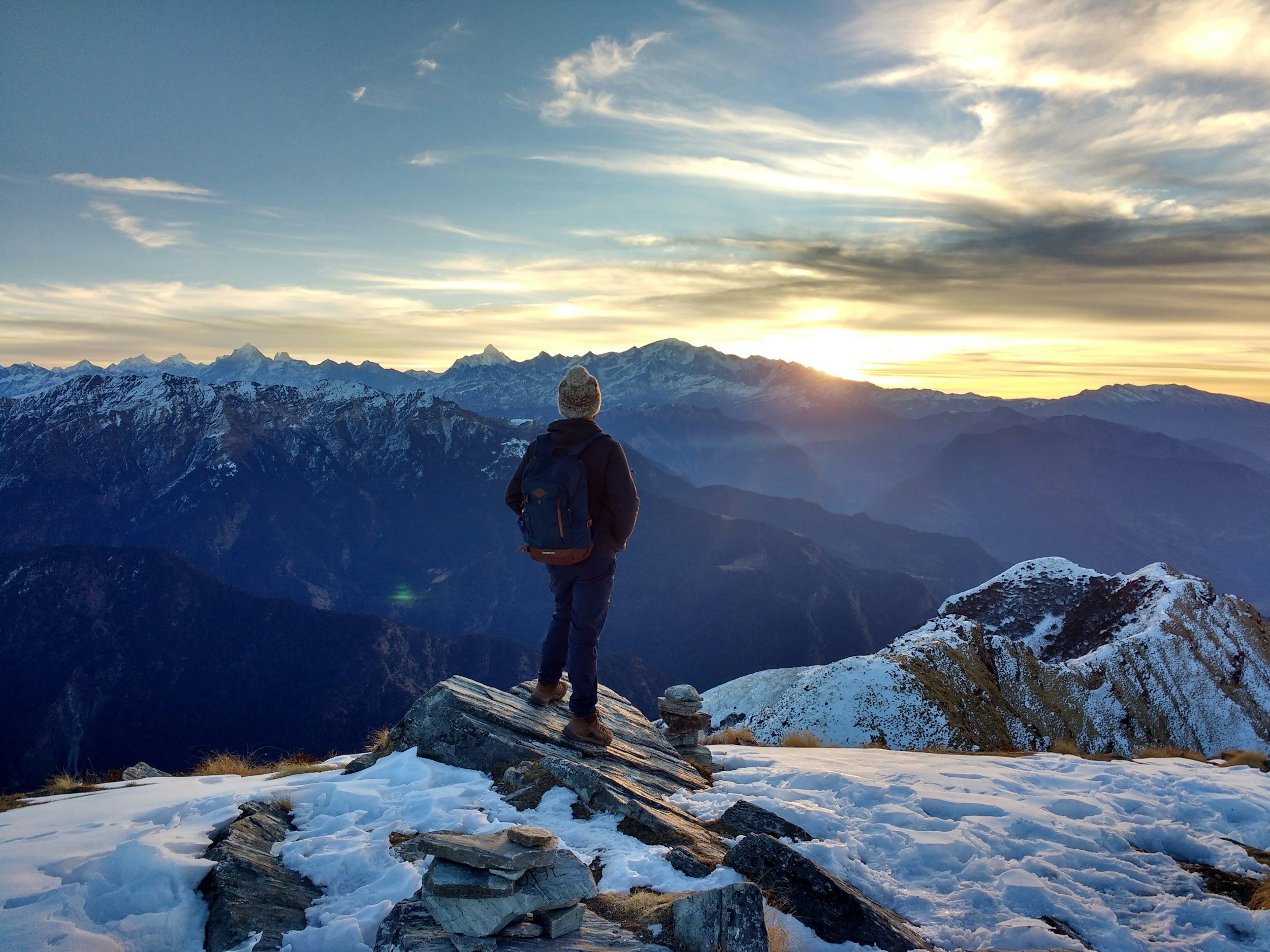 Une personne solitaire en vêtements d'hiver se tient sur un rocher au sommet d'une montagne enneigée, regardant un paysage de montagnes majestueuses sous un ciel coloré au coucher du soleil ou à l'aube.
