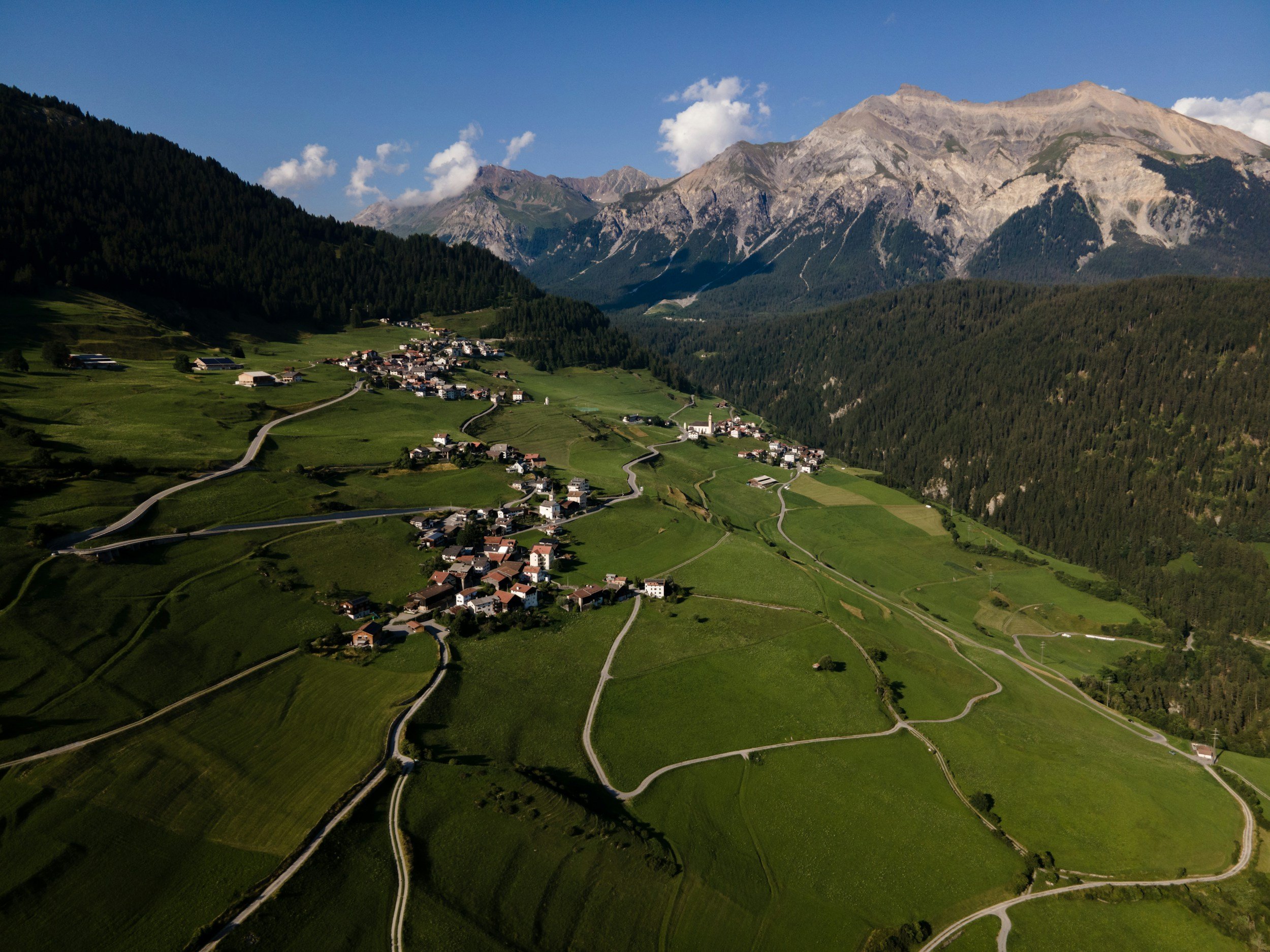 Vallée verdoyante avec des Maisons, des routes sinueuses et des montagnes en arrière-plan sous un ciel bleu avec quelques nuages.