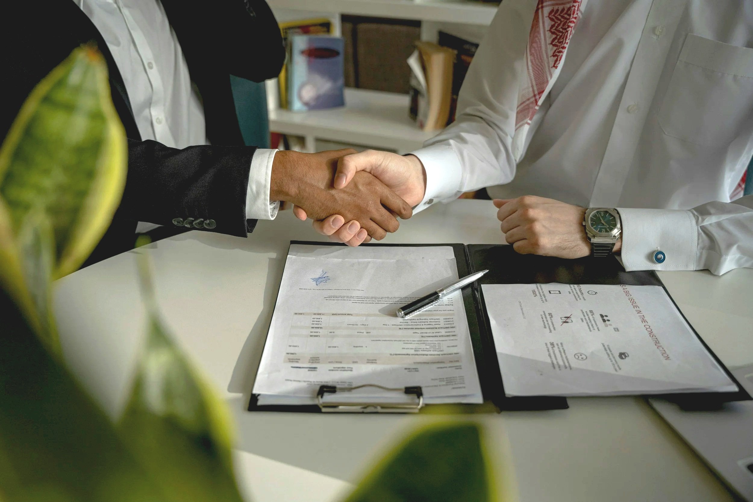 Deux personnes se serrent la main au-dessus d'une table avec des documents et un stylo, dans un environnement de bureau, avec une plante verte floue au premier plan.
