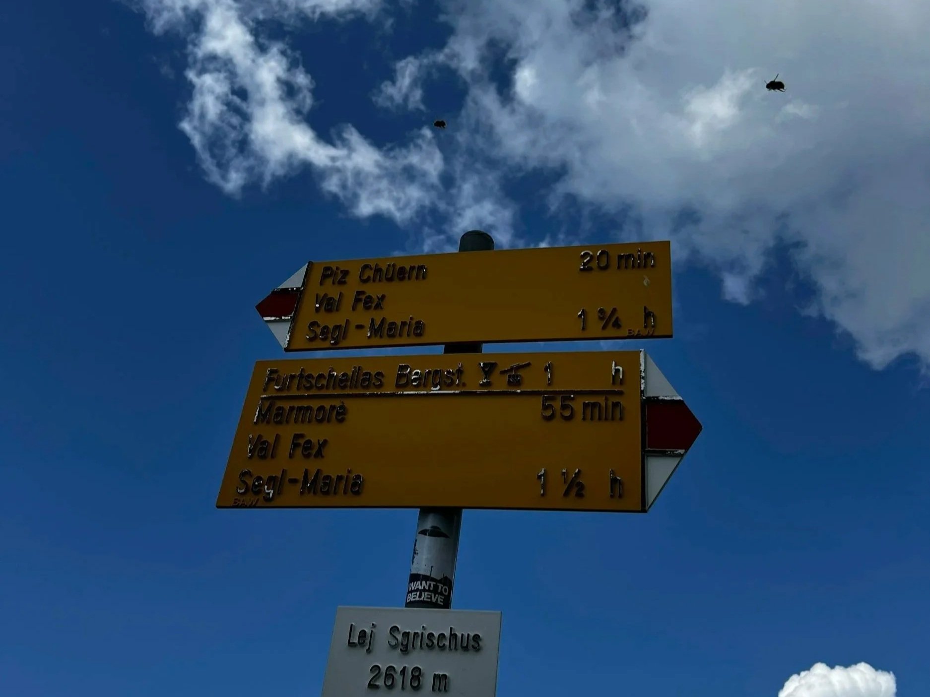 Panneau indicateur de direction en montagne avec le ciel bleu en arrière-plan, indiquant les distances à des endroits comme le Pla Chur, Val Fex et Seghil-Marie.