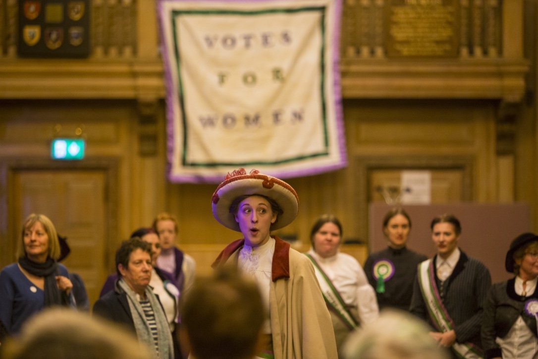 We Are Outlaws, 2018. A woman in Edwardian dress addresses a crowd, a large banner reading 'Votes for Women' behind her. 