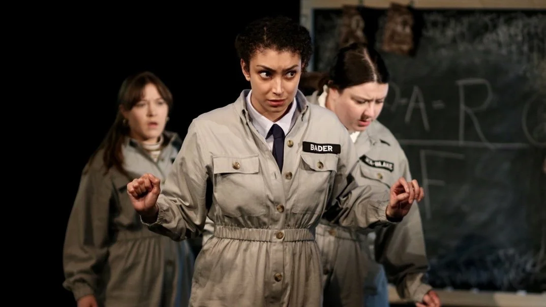 Unsung, 2019. Three women look concerned, all wearing identical boiler suits sporting the surnames 'Bader', 'Jex-Blake' and 'Dunbar'.