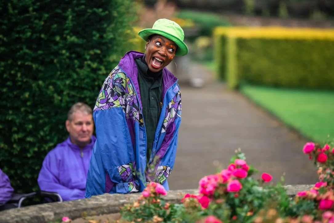 A Midsummer Night's Dream, 2021. A woman wearing festival paint and a jazzy raincoat stands in a rose garden with her eyes and mouth wide.