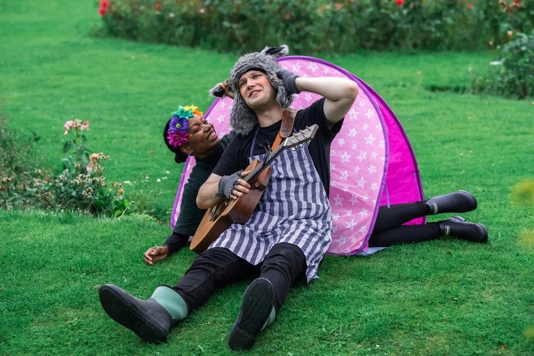A Midsummer Night's Dream, 2021. A man in a donkey hat, holding a guitar, enjoys a romantic moment with a woman wearing a wreath of flowers, sitting in front of a pink tent.