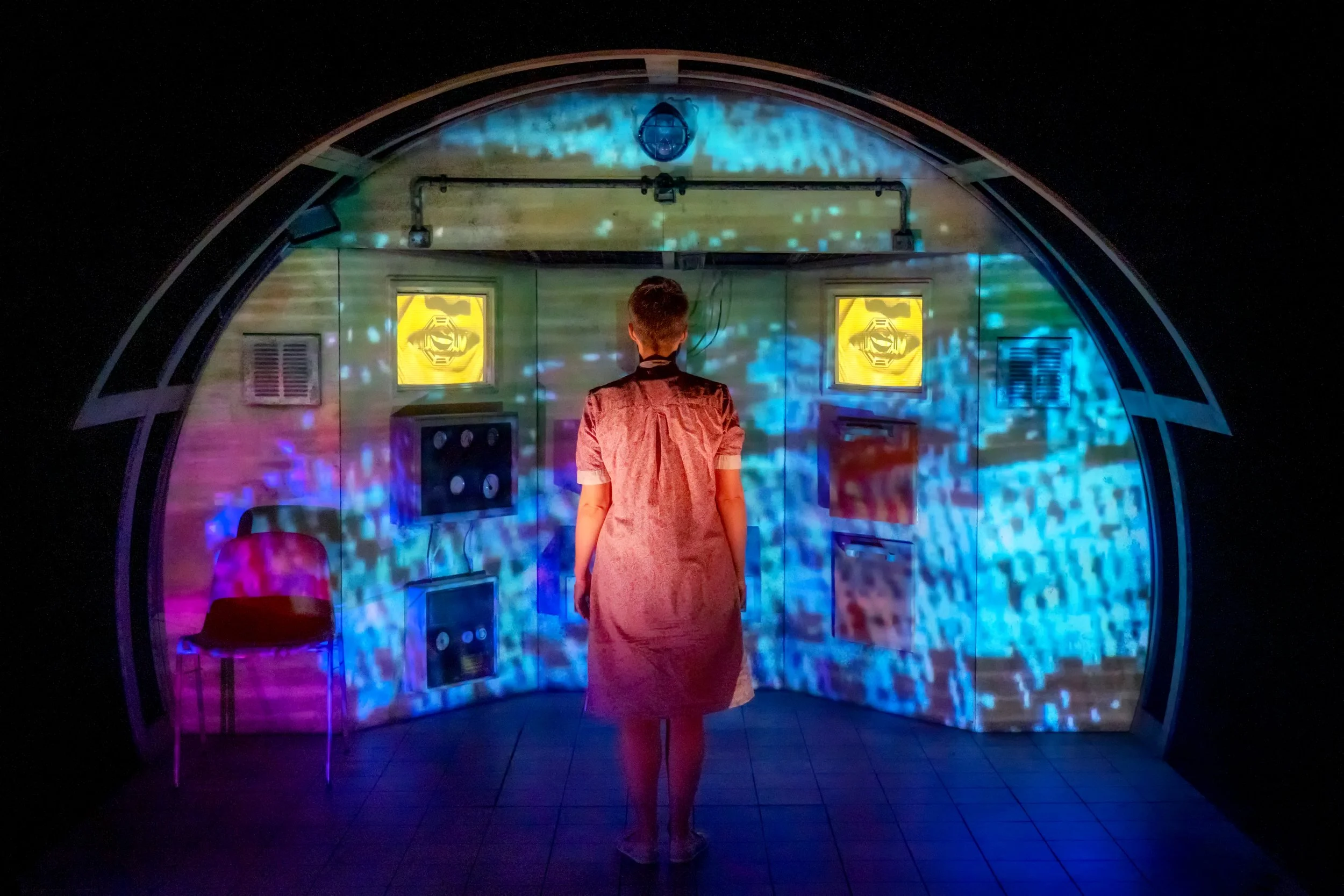 Smile Club, 2020. A woman stands in a night dress, with her back to the camera, with a dystopian room behind her.