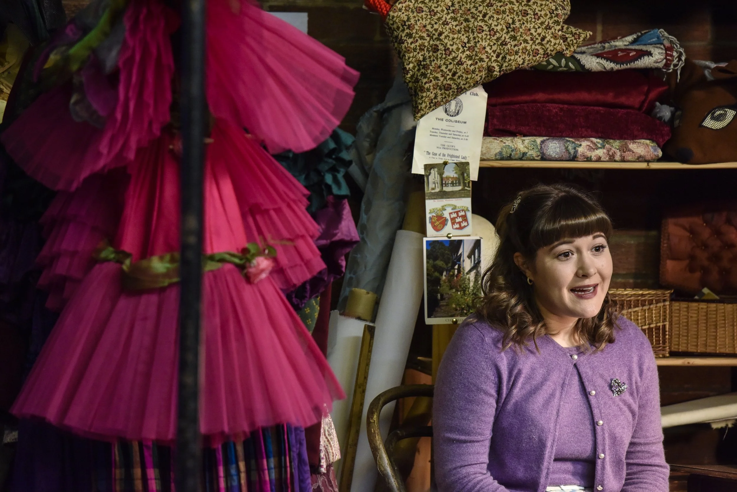 Whodunnit at the Coliseum?, 2021. A young woman sits in a costume store, speaking to someone off-camera.