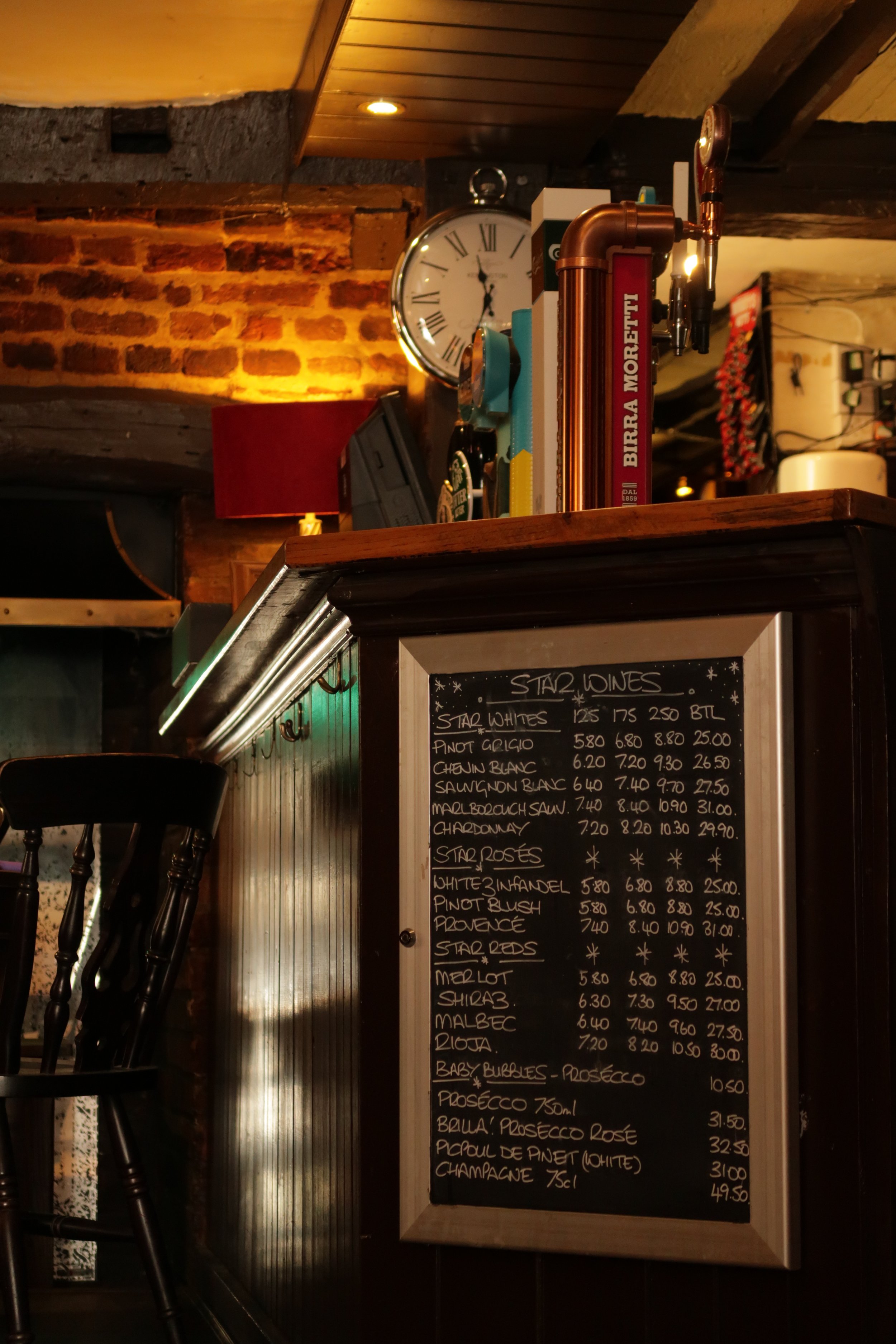 A chalkboard menu listing various wine options and prices in a cozy bar or restaurant with wooden accents, a brick wall, and warm lighting. There's a bar counter with bar stools in the foreground, and in the background, a clock on the wall.