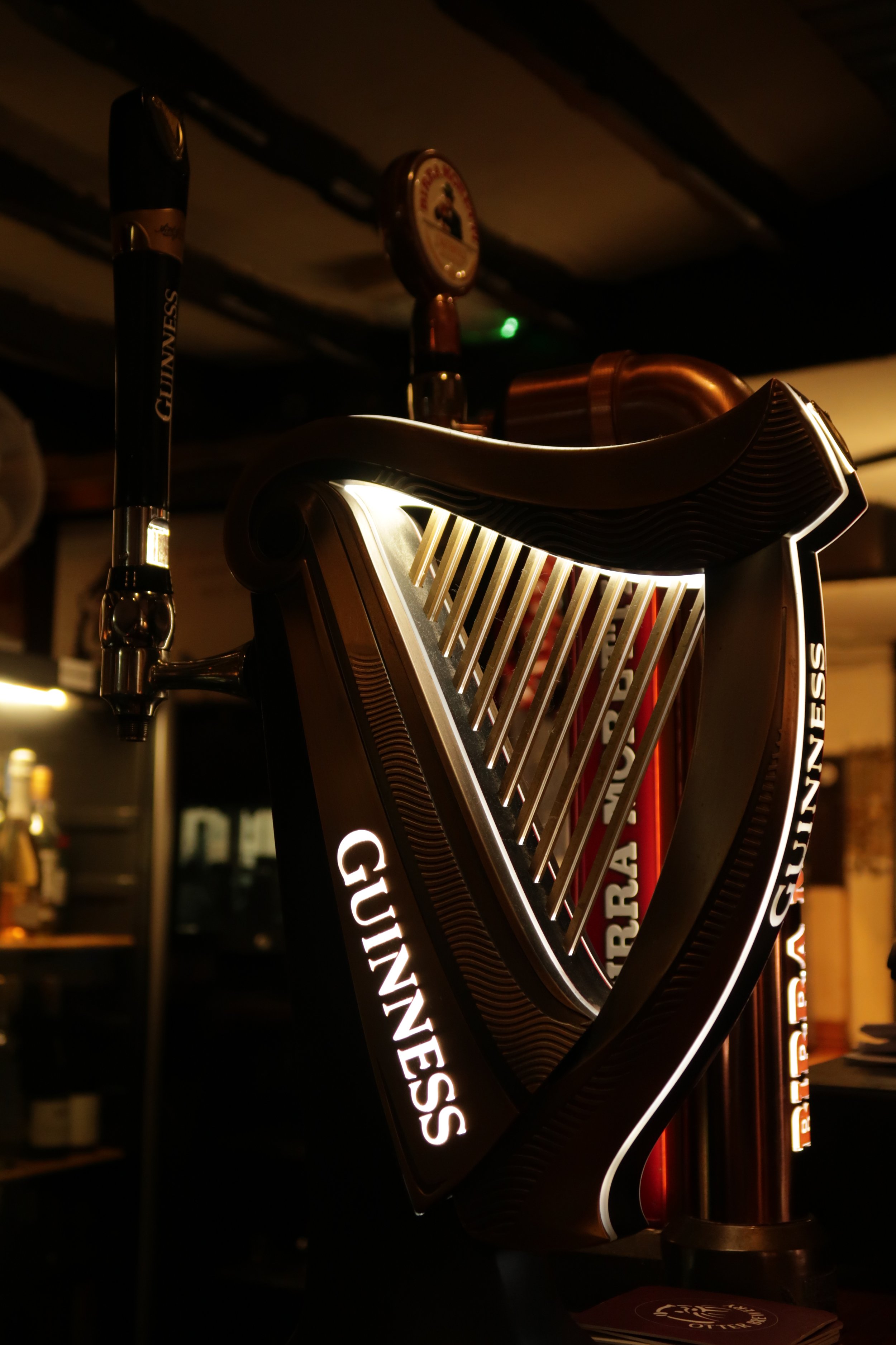 A wall-mounted Guinness beer tap with illuminated branding, attached to copper pipes, in a dimly lit bar setting.