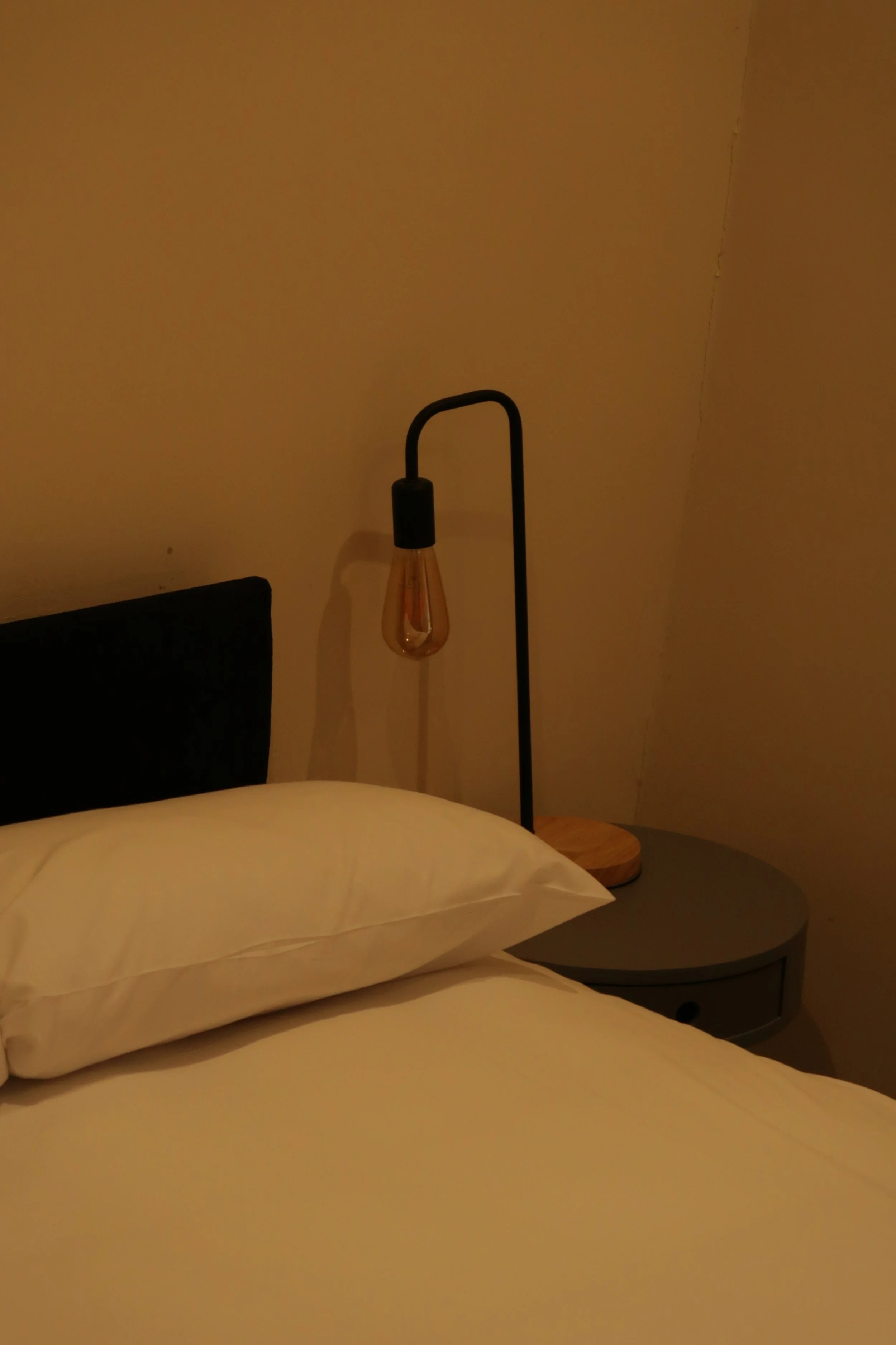 A bedroom scene with a bed, white pillow, bedside table, and a floor lamp with an exposed light bulb against a plain yellow wall, a pub in ringwood, Dorset