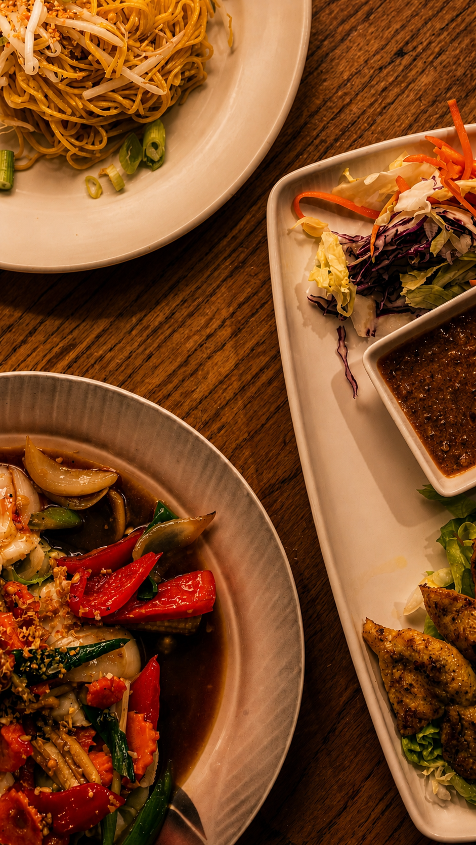 Close-up of plates of Asian-style dishes, including stir-fried vegetables, noodles, salad, and dipping sauces on a wooden table.