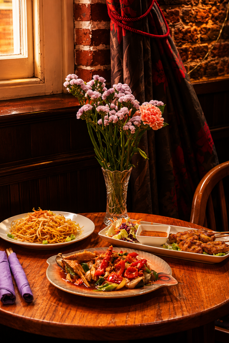 A cozy restaurant table with a vase of pink and orange flowers, a plate of stir-fried vegetables, a plate of noodles with vegetables, and a dish of grilled chicken skewers with salad and dipping sauce, near a window with dark curtains and brick wall.
