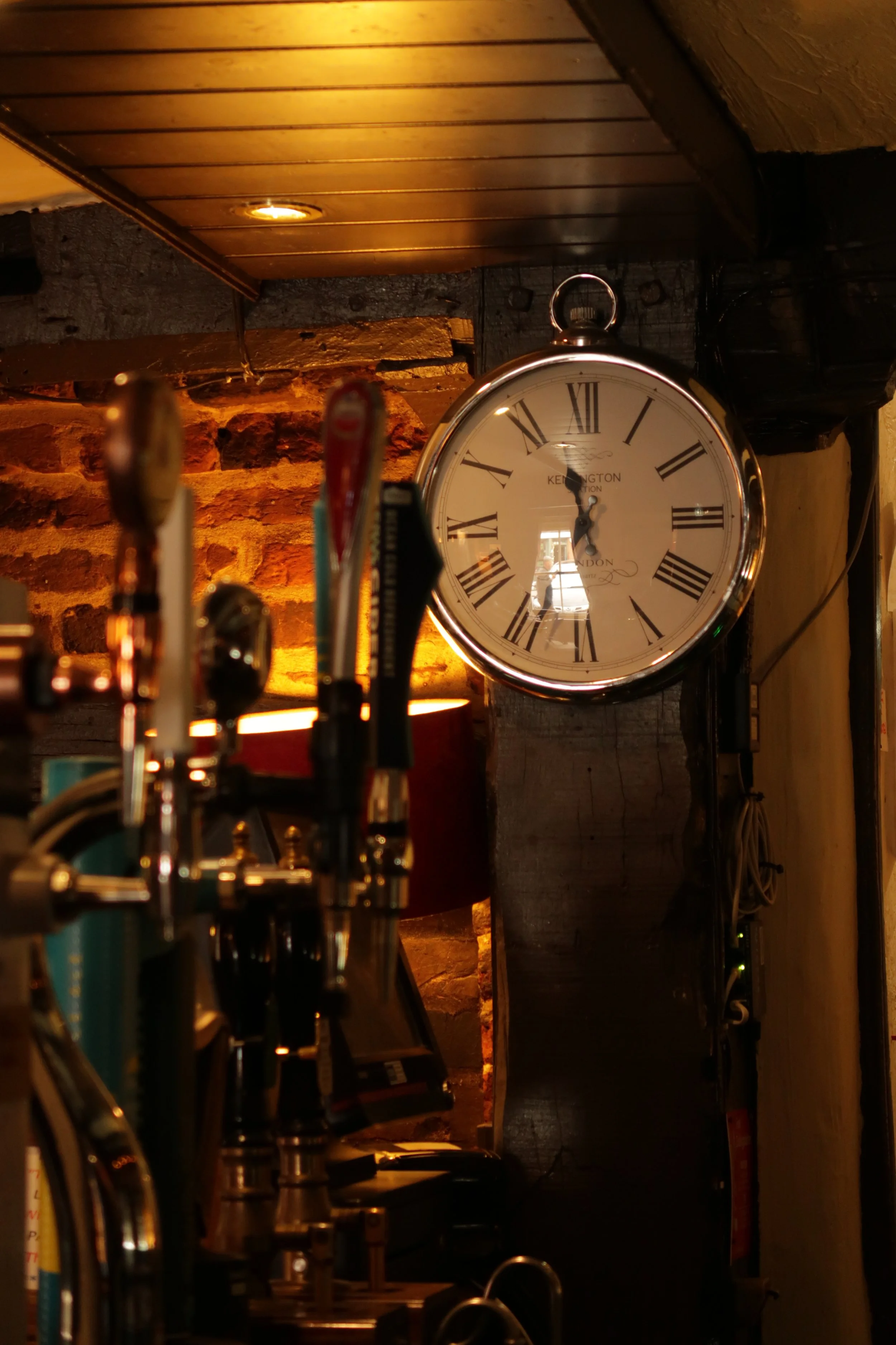 A large clock with Roman numerals on the wall behind a row of beer taps in a bar or restaurant.