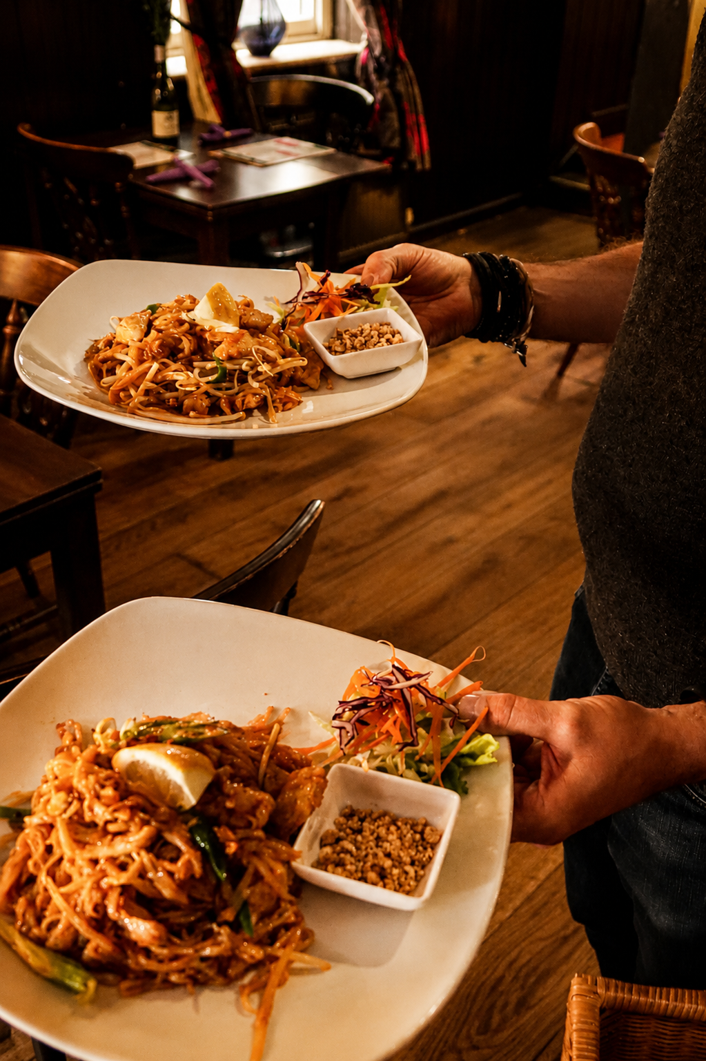 Two people holding serving plates of Chinese stir-fried noodles with vegetables, lemon wedges, a small bowl of crushed peanuts, and a side salad, inside a cozy restaurant with wooden furniture, a pub in ringwood, Dorset