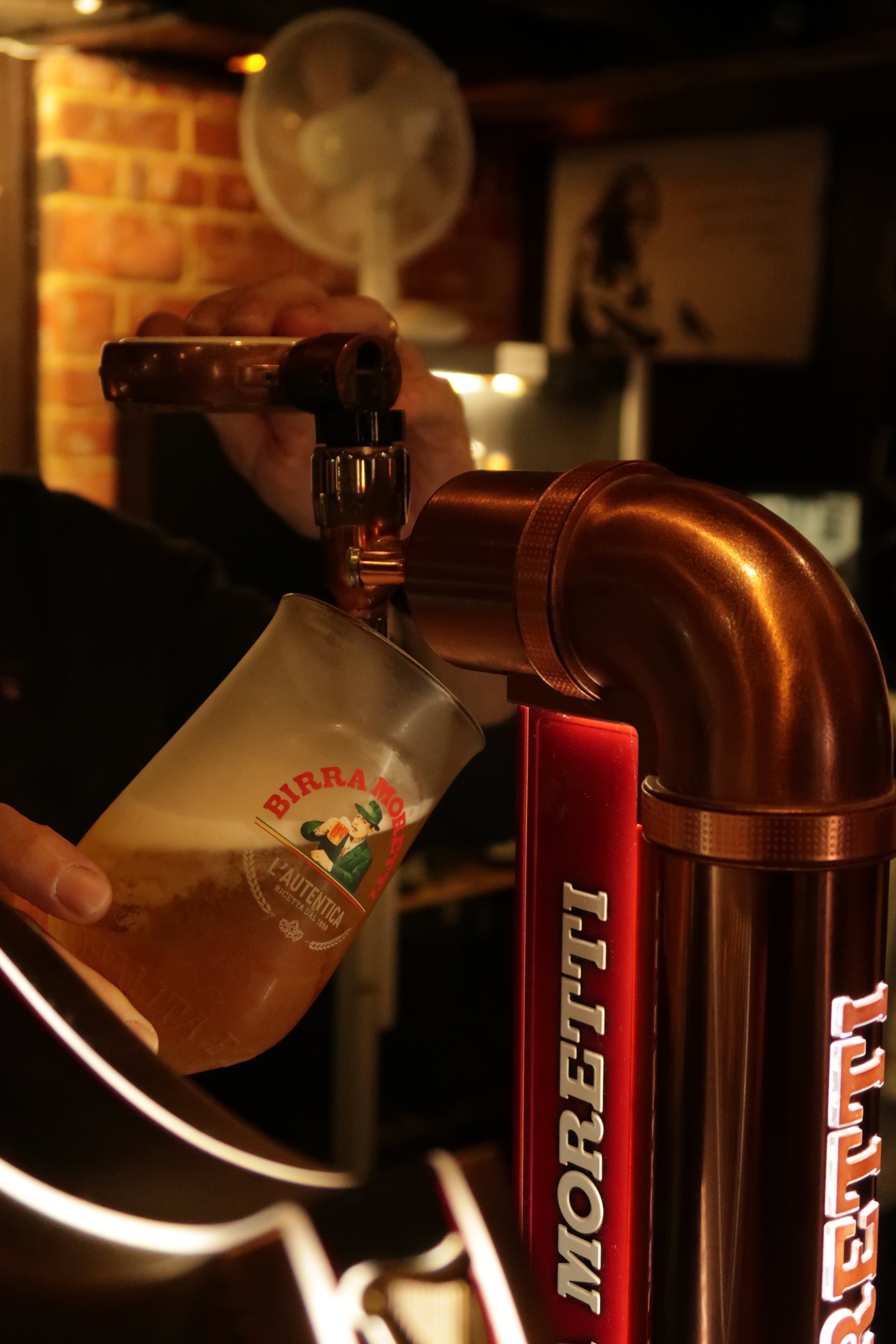 Close-up of a hand pouring BIRRA MORETTI beer into a glass at a bar, with a copper beer tap, a fan, a brick wall, and framed artwork in the background, a pub in ringwood, Dorset