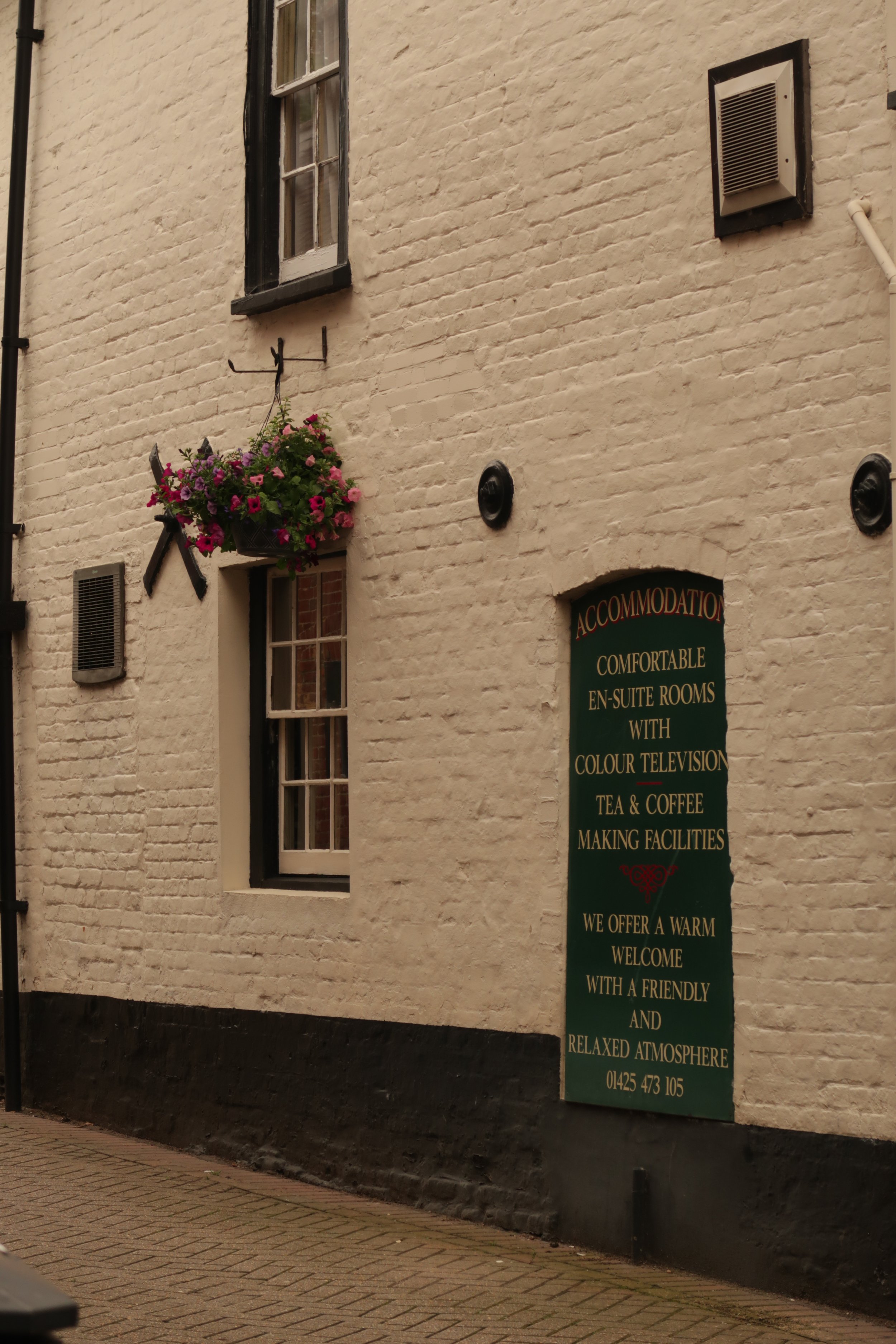The image shows a white brick building with a window and a hanging flower basket containing pink and purple flowers. To the right, there is a green signboard with gold writing advertising accommodations, en-suite rooms, tea and coffee facilities, and