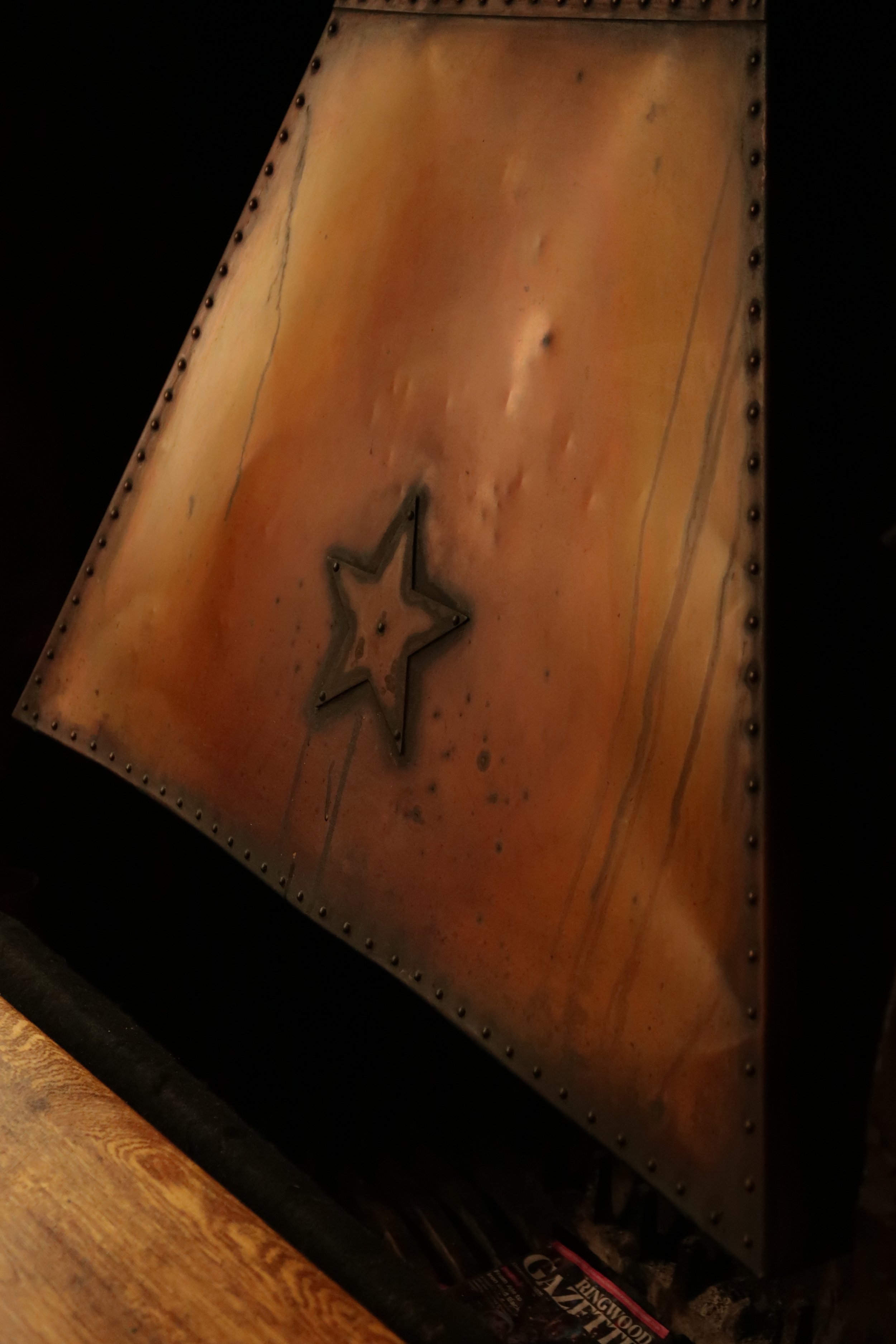 A close-up image of a vintage metal sign with a star design, featuring a warm, orange-brown color with visible wear and rivets along the edges, placed on a dark surface with part of a wooden table in the foreground and a magazine underneath.