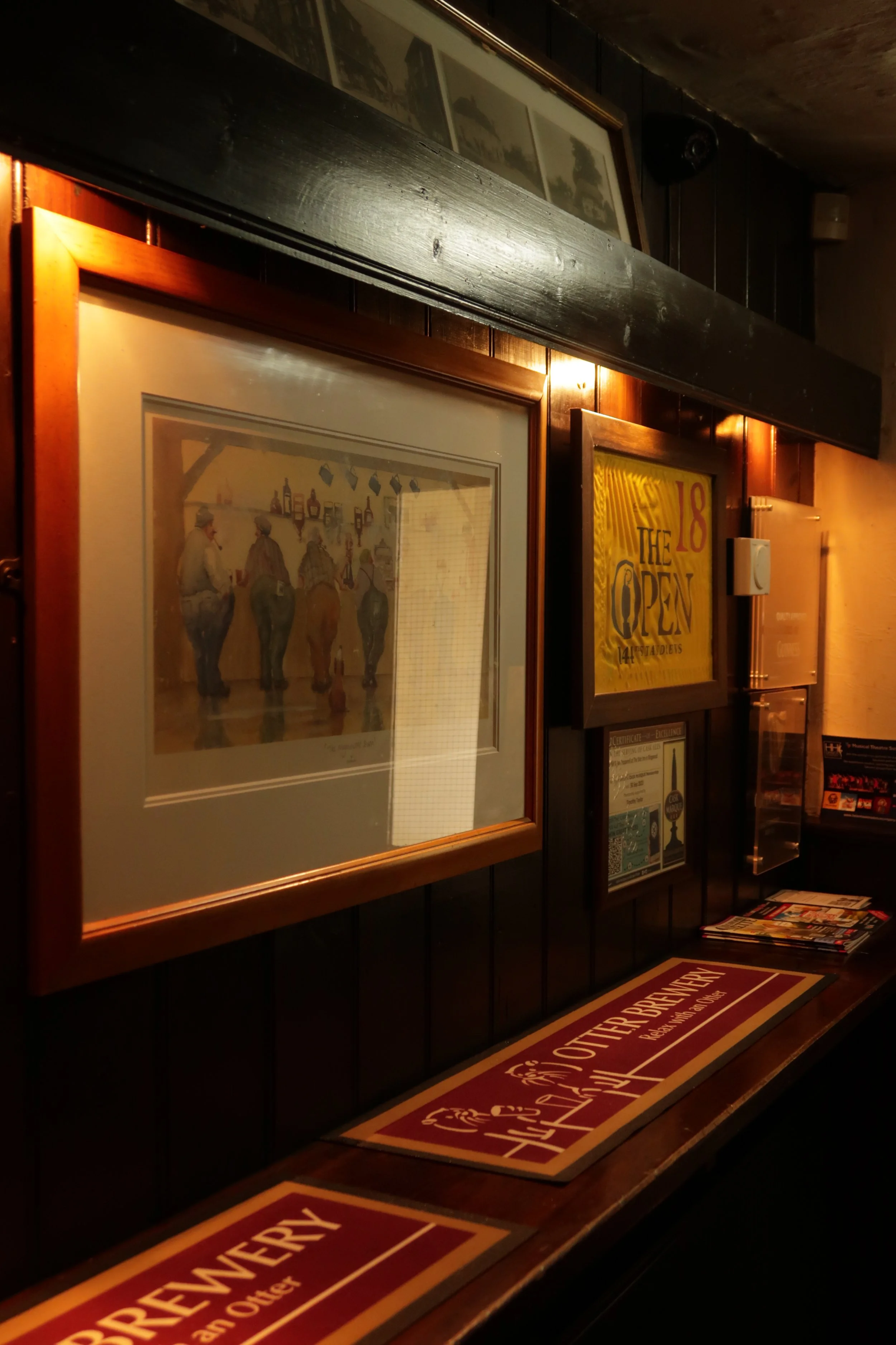 Interior of a pub or bar with decorated wooden walls, framed pictures, and various signs including a yellow sign that says 'The 18th Open' and a red and yellow sign on the floor that says 'Deli's Osterbry.