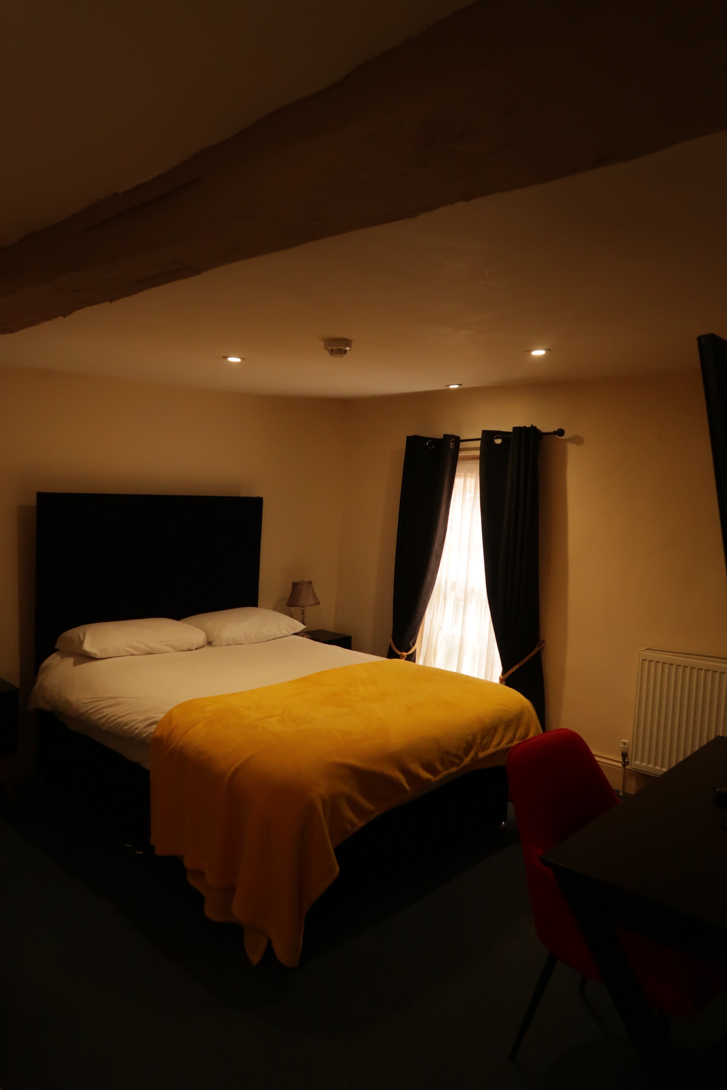 Dimly lit bedroom with a bed covered in a yellow blanket, two pillows, small bedside table with lamp, window with dark curtains, and a red chair in the foreground, a pub in ringwood, Dorset