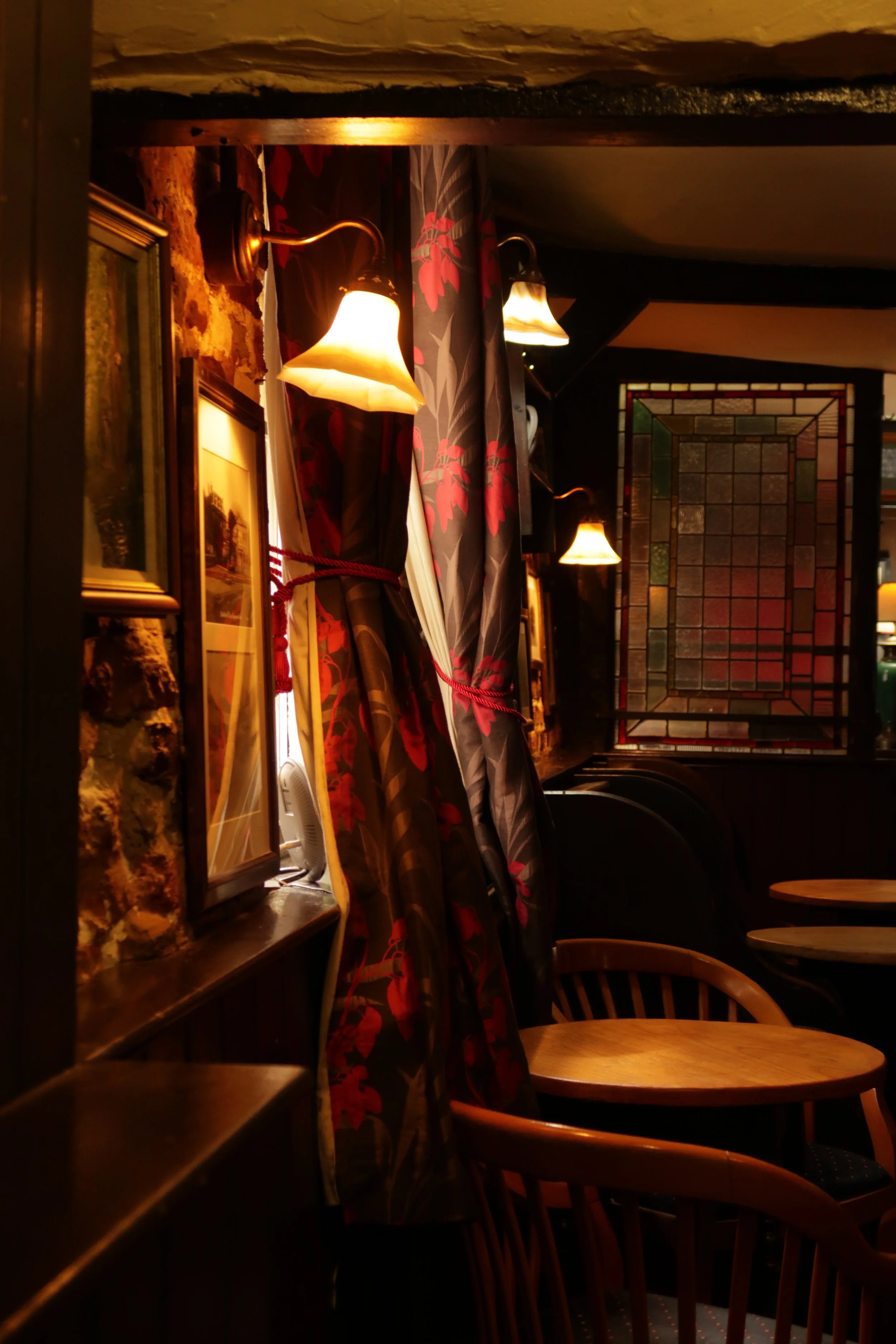 Cozy indoor restaurant corner with wooden tables, chairs, floral curtains, wall lamps, framed pictures, and stained glass window.