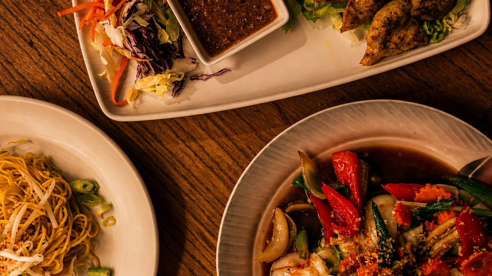 Close-up of Asian food dishes on a wooden table, including noodle stir-fry with bean sprouts, a salad with shredded vegetables and dipping sauce, and a stir-fried vegetable and meat dish with bell peppers and onions.