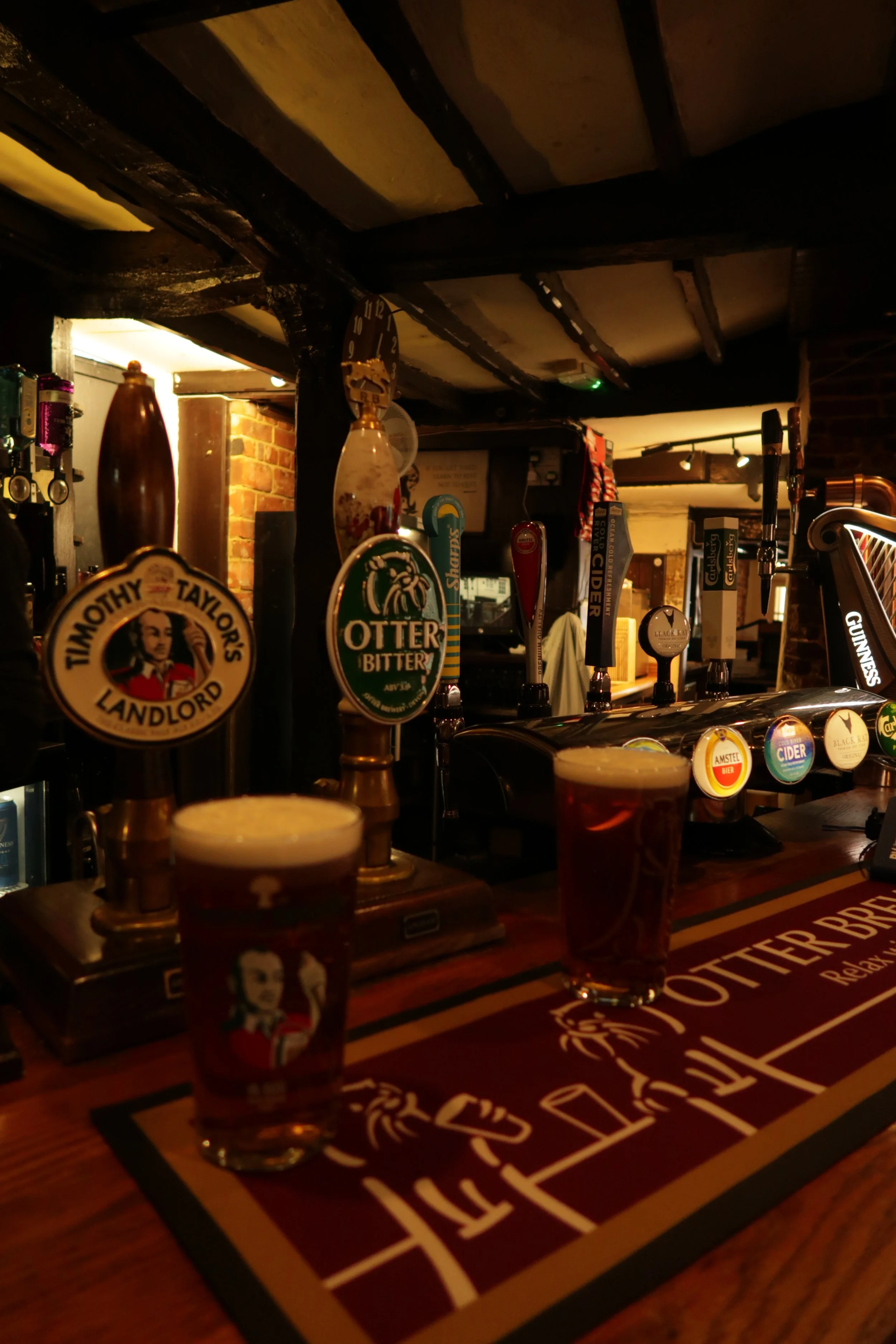 A bar with draft beer taps and pints of beer on the counter, illuminated with warm lighting, with exposed brick and wooden beams overhead, a pub in ringwood, Dorset