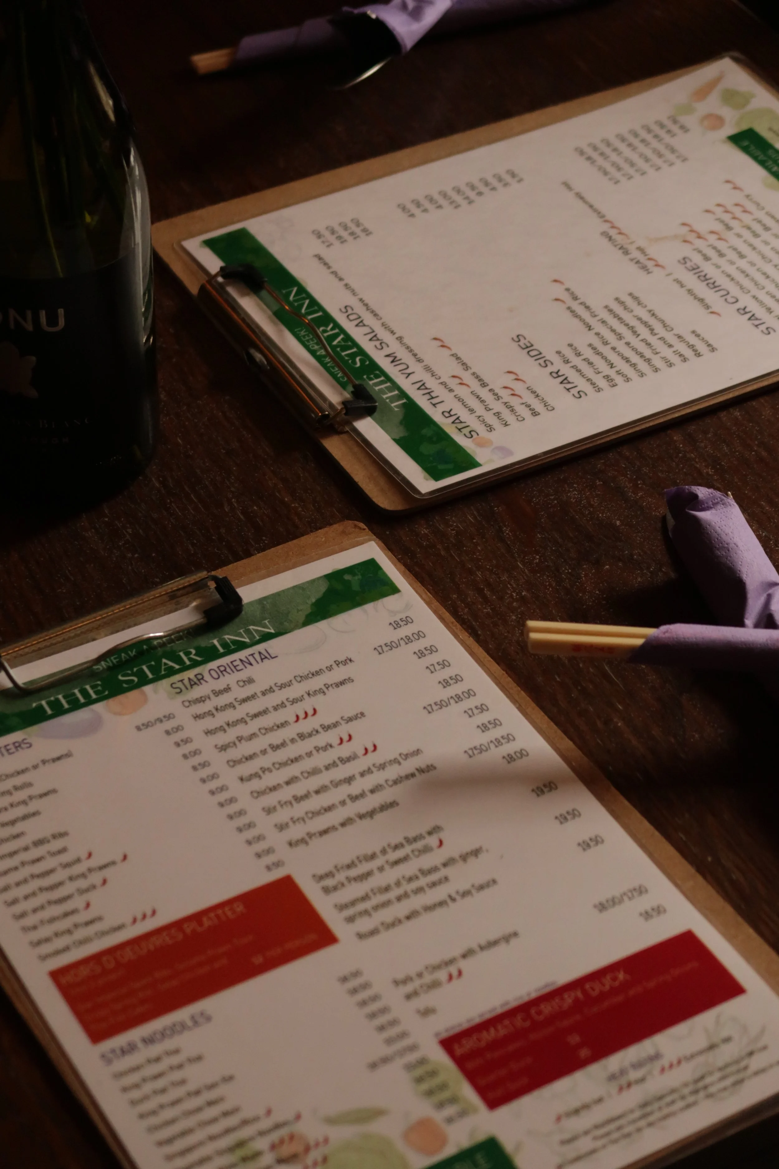 A menu on a clipboard with various food and drink options, set on a dark wooden table, with a purple cloth napkin and chopsticks nearby.