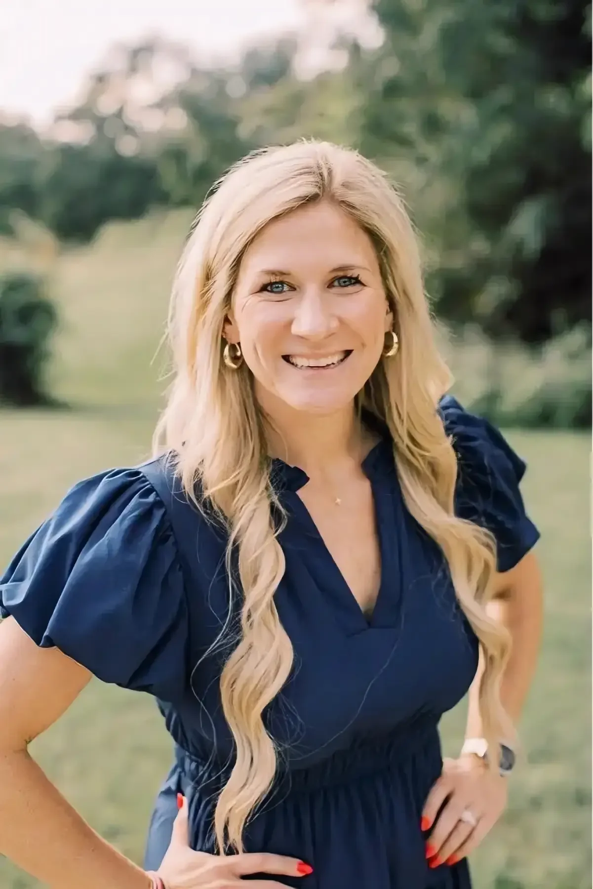 A smiling woman with long blonde hair, wearing a navy blue dress with puffed sleeves, standing outdoors with green trees in the background.