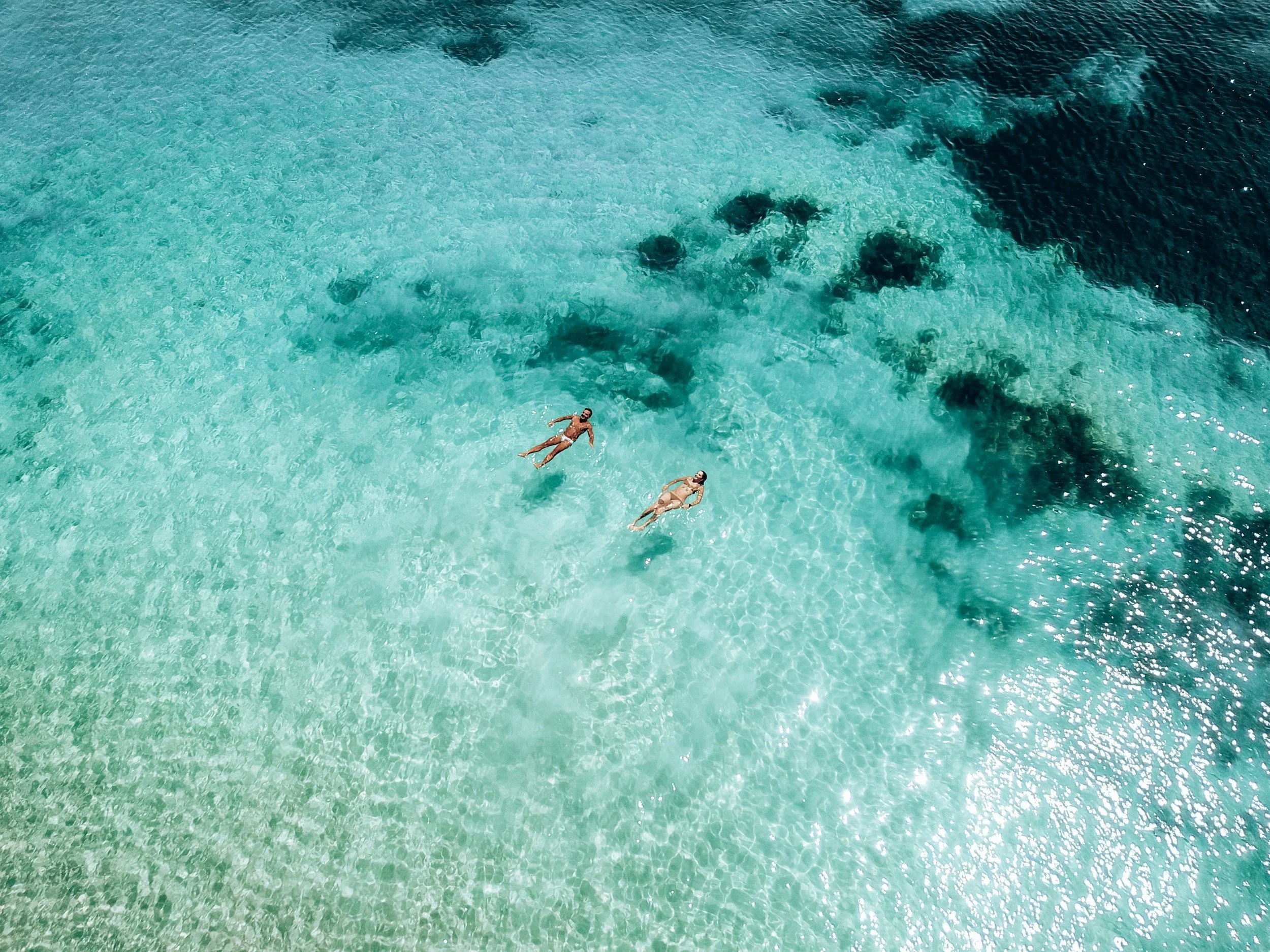 Aerial view of two people floating in clear turquoise water near some dark patches of coral or rocks.