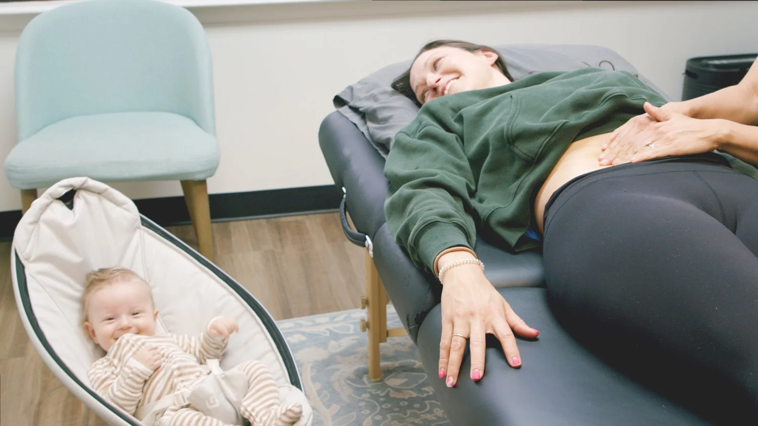 A postpartum recovery client receives treatment while her infant smiles nearby in our kid-friendly treatment office