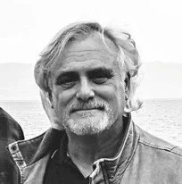 The author, Ronald J. Pauley, with gray hair and a beard, wearing a denim jacket, is outdoors in the mountains of Montana.