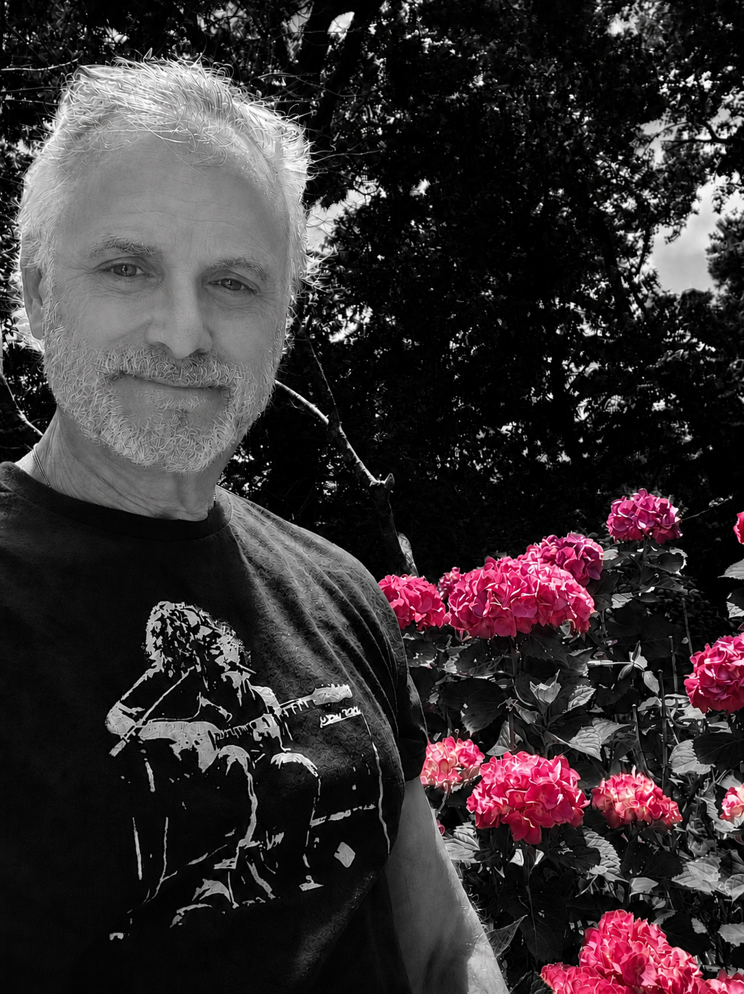 Ron, with white hair and a beard, smiling, standing outdoors near pink hydrangea flowers and greenery.