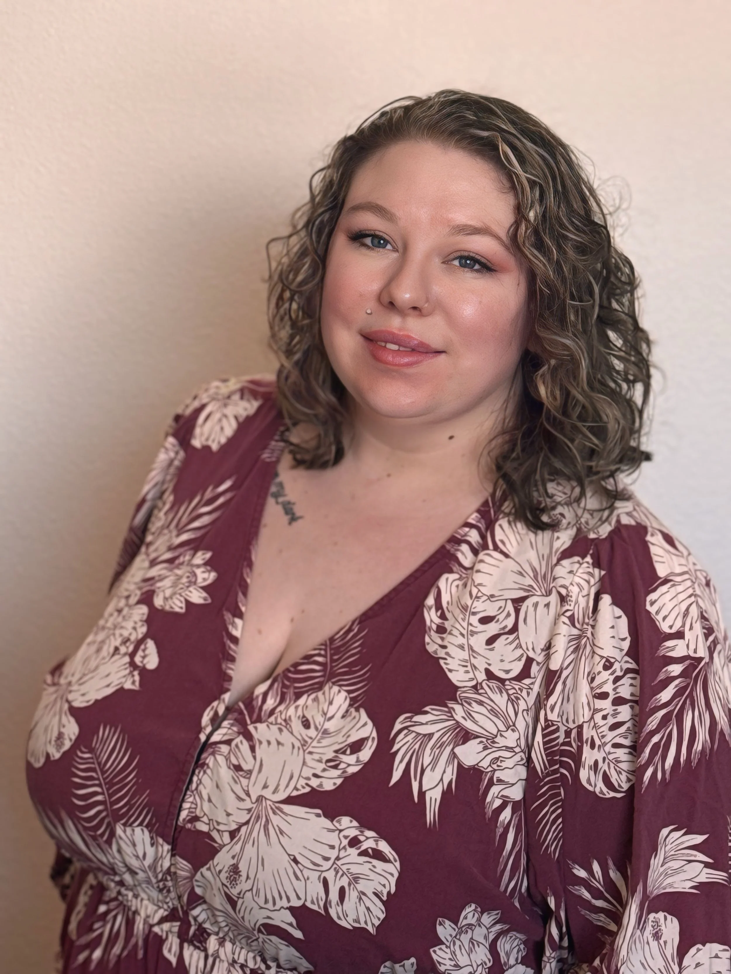 Sarah Gajewski with curly hair and blue eyes smiling slightly, wearing a maroon dress with a beige floral pattern, standing against a beige wall.